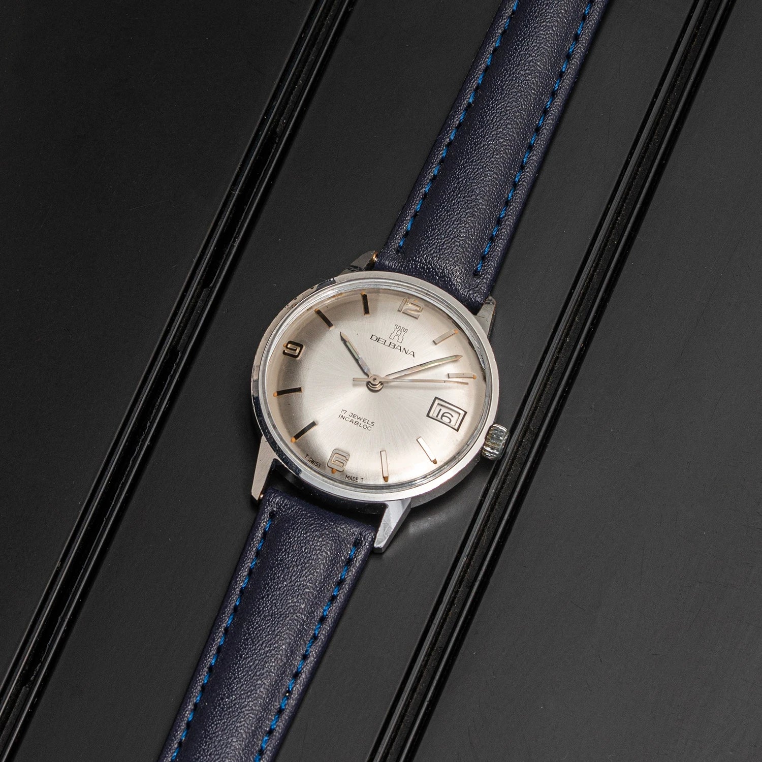 A vintage-style wristwatch Delbana, Cal. AS 1951 with a silver-toned oval case, cream dial, and dark blue leather strap with bright blue stitching, photographed on a dark textured background.