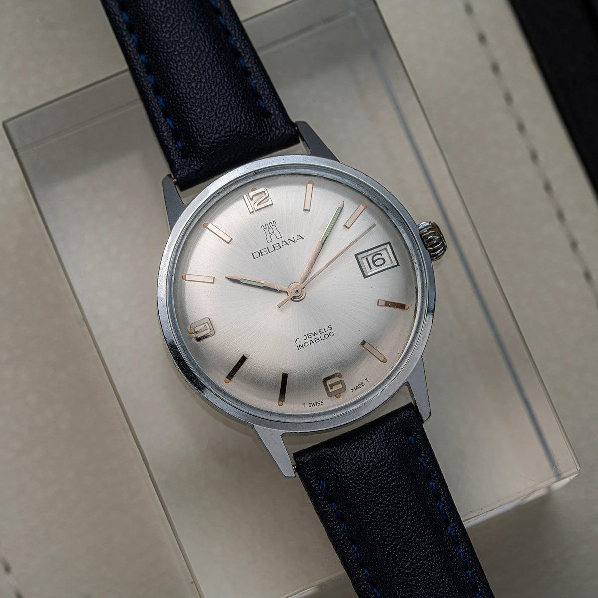 A vintage Delbana, Cal. AS 1951 analog watch with a silver dial, rose gold-tone markers and hands, and a dark blue leather strap, resting on its beige presentation box.