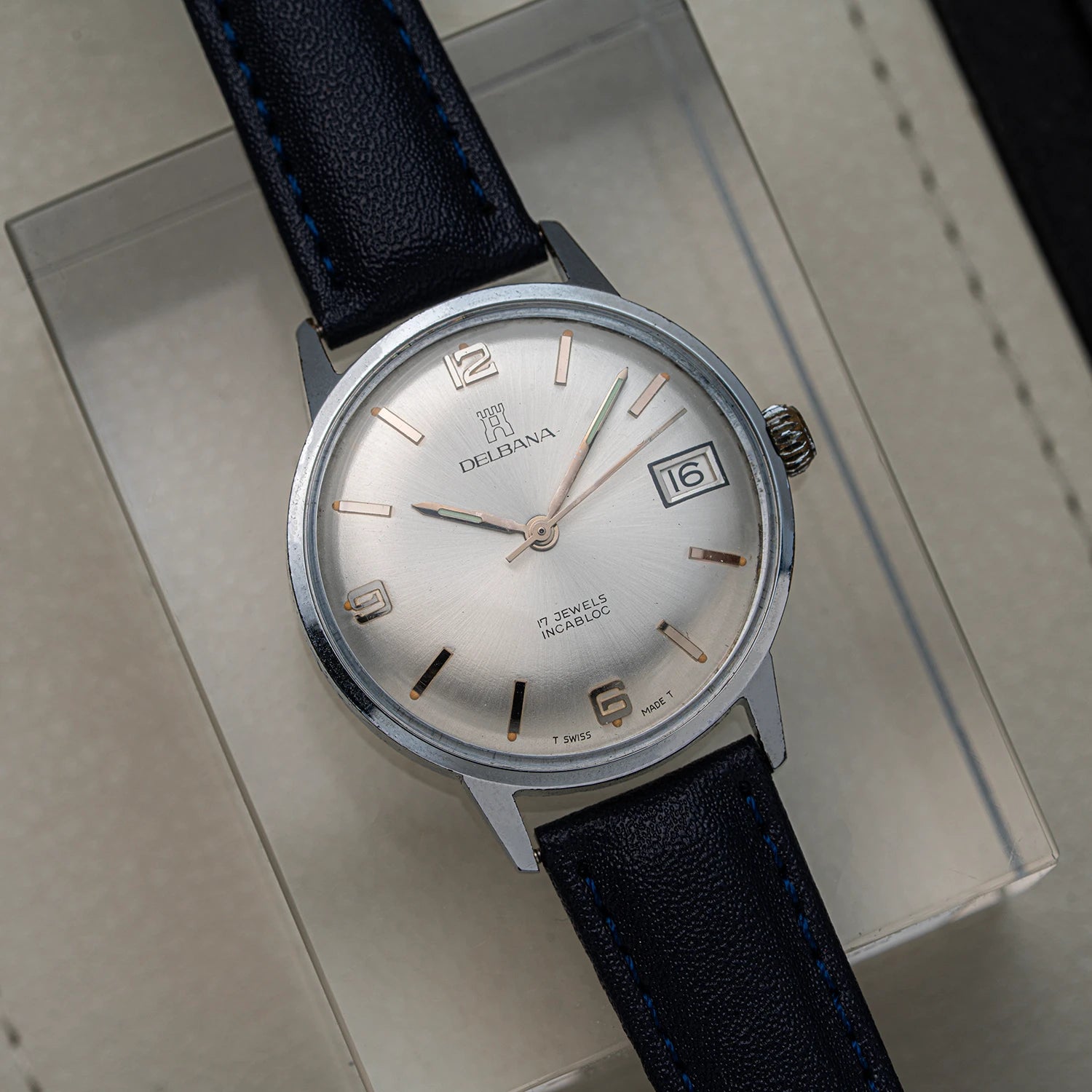 A vintage Delbana, Cal. AS 1951 analog watch with a silver dial, rose gold-tone markers and hands, and a dark blue leather strap, resting on its beige presentation box.