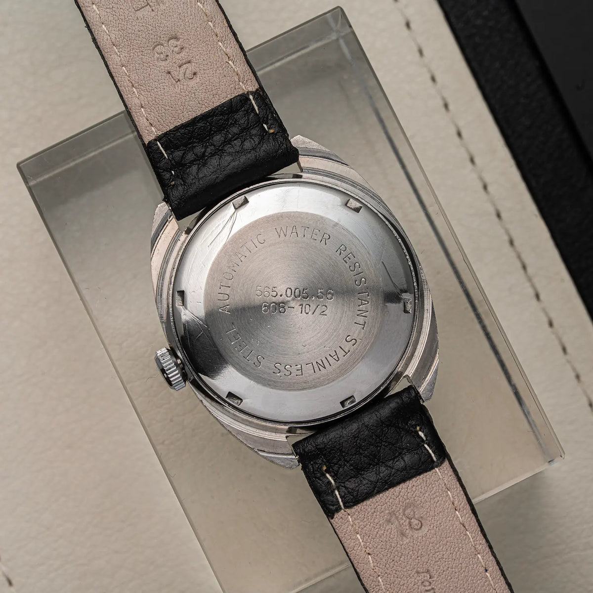 Close-up of the back of a wristwatch Doxa Neuchatel, Ref. 565.005.56, Cal. AS 5206, Men’s Automaticwith a stainless steel case and a light beige leather strap with black stitching, resting on a clear display block.
