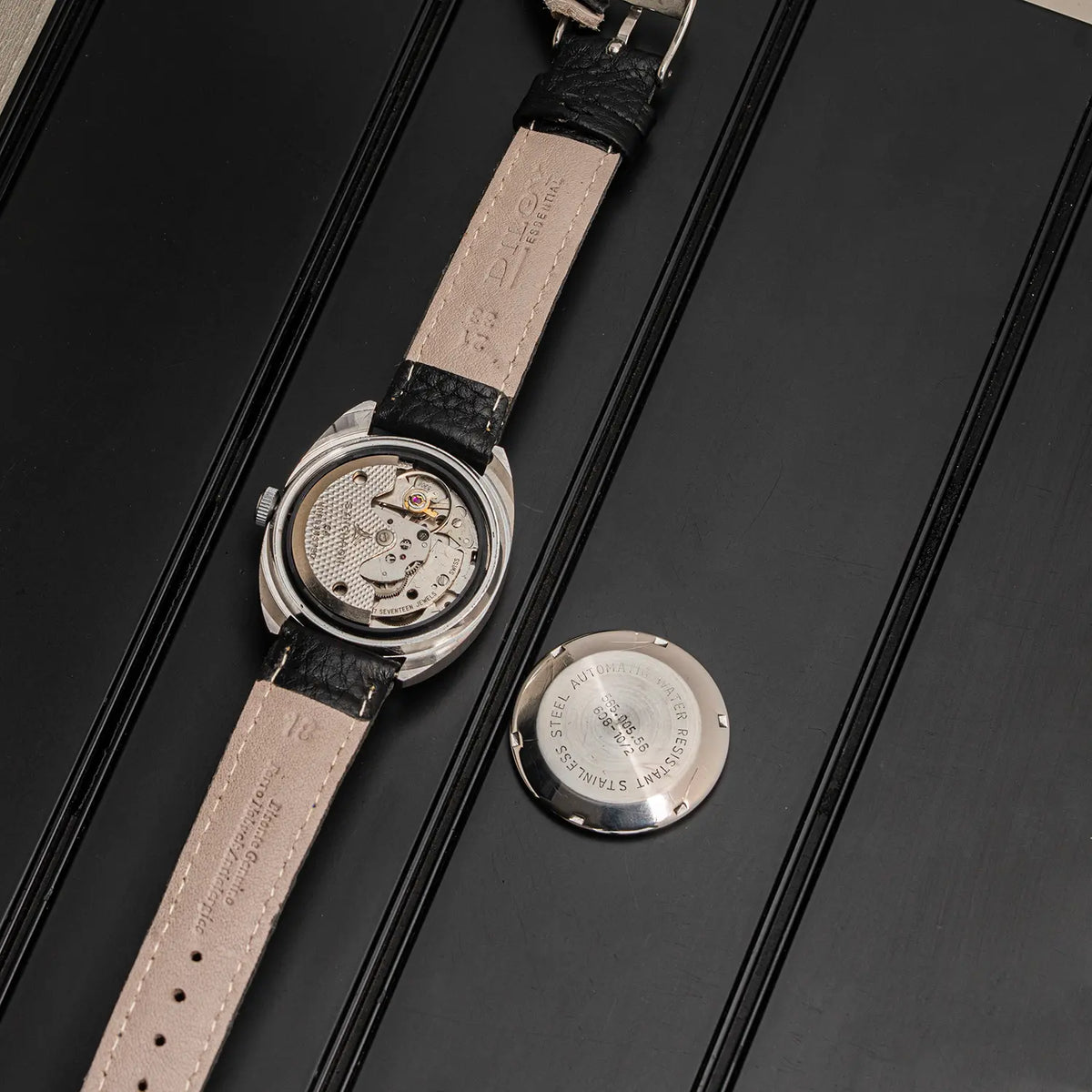 A vintage-style mechanical watch Doxa Neuchatel, Ref. 565.005.56, Cal. AS 5206, Men’s Automatic with a silver-toned case, showing the exposed automatic movement through a transparent back, laid diagonally on a black textured surface next to its circular metal back cover.