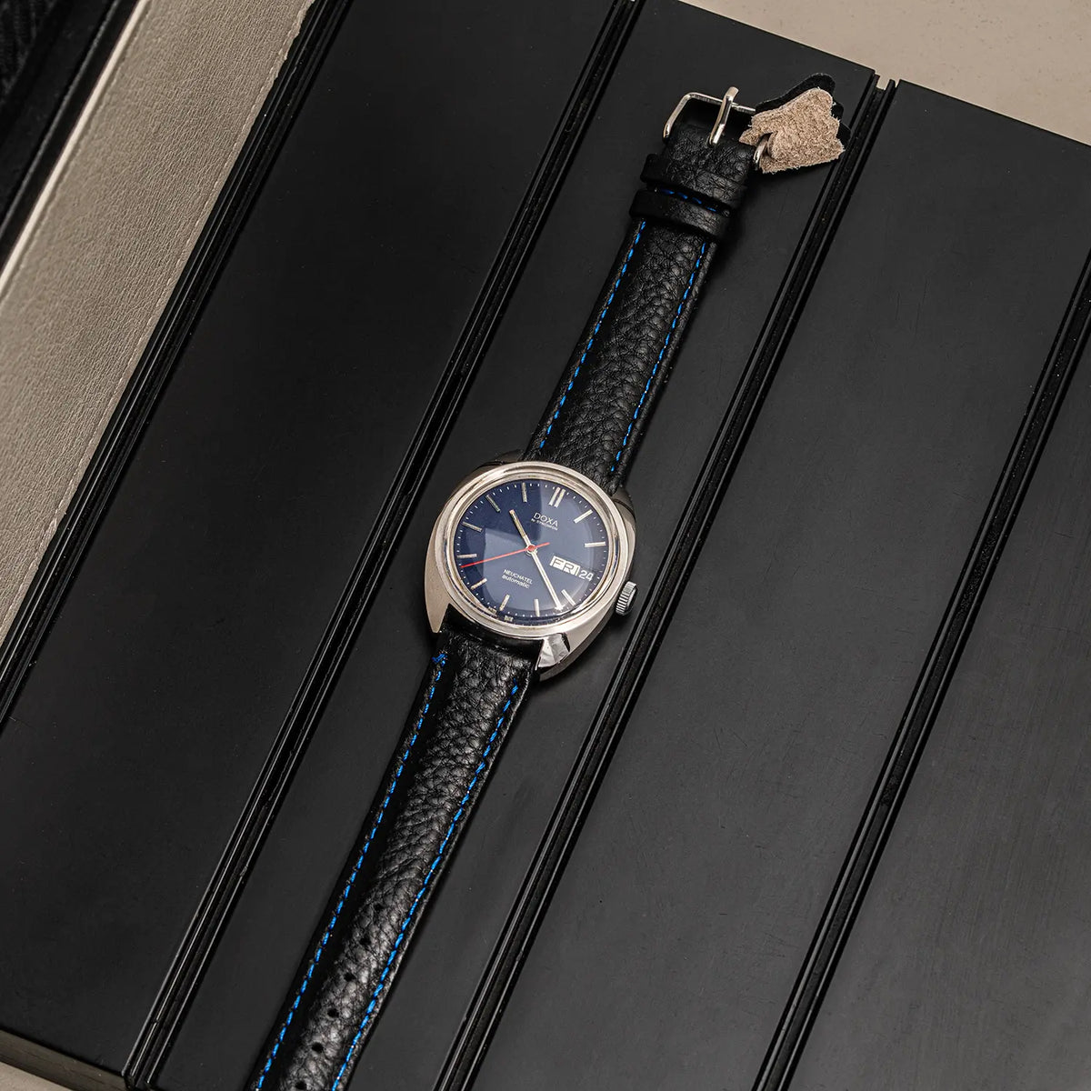 A wristwatch Doxa Neuchatel, Ref. 565.005.56, Cal. AS 5206, Men’s Automatic with a blue dial and a textured black leather strap with blue stitching, displayed diagonally on a black textured surface.