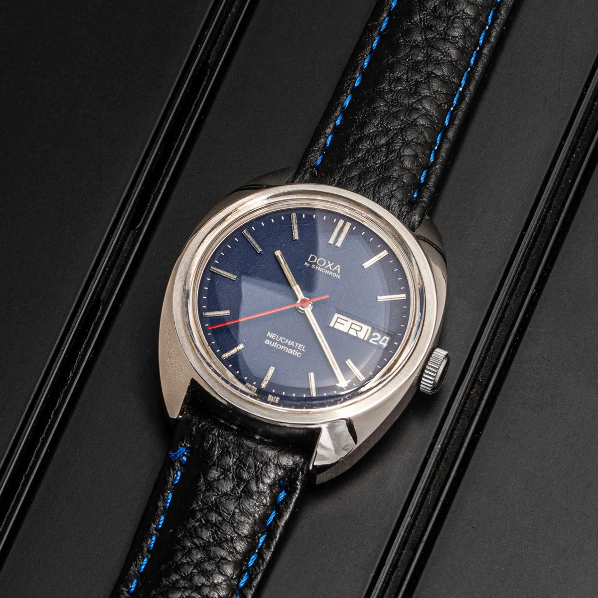 A vintage-style Doxa Neuchatel, Ref. 565.005.56, Cal. AS 5206, Men’s Automatic wristwatch with a round silver-tone case, dark blue dial, and black textured leather strap with bright blue stitching, photographed on a dark background.