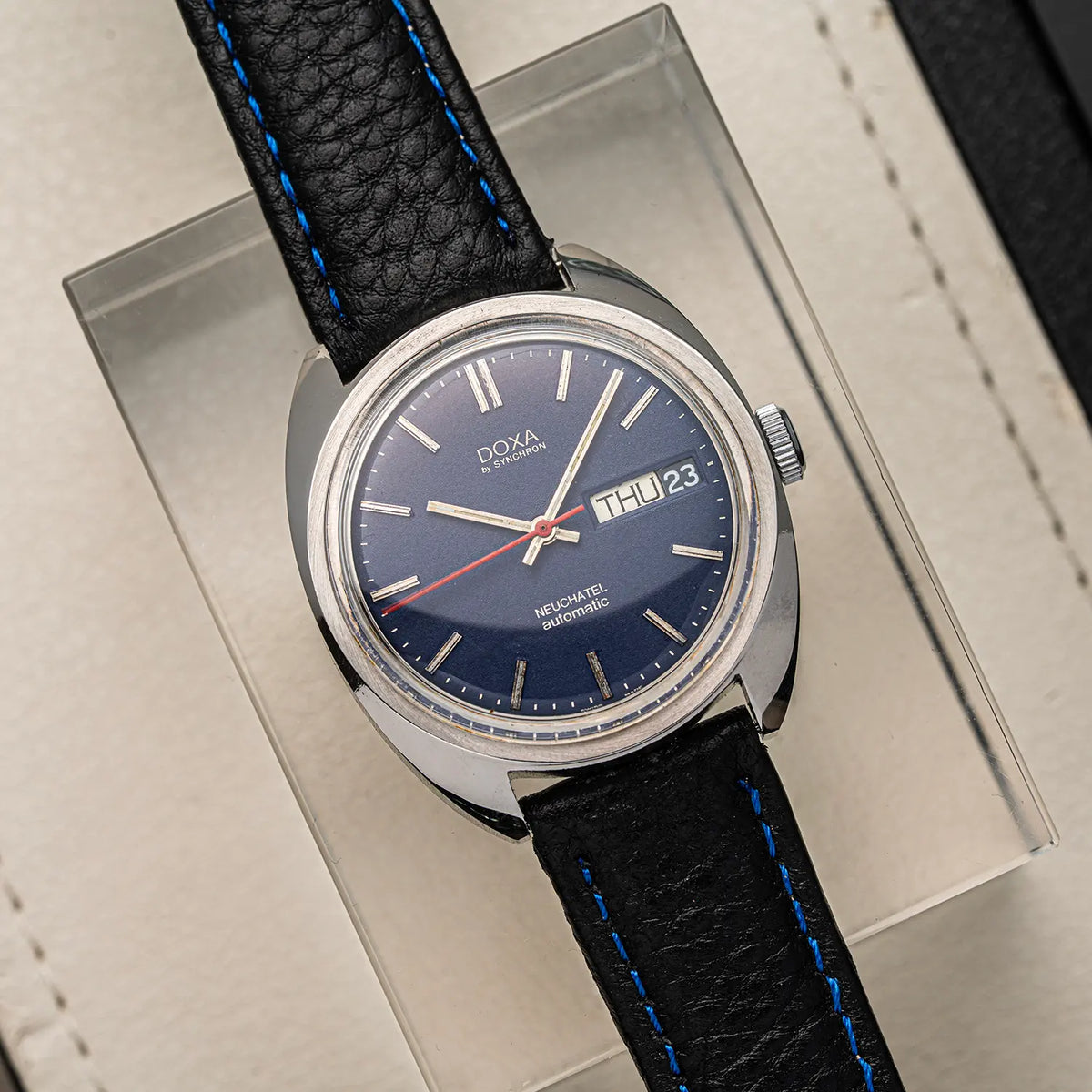A vintage-style wristwatch Doxa Neuchatel, Ref. 565.005.56, Cal. AS 5206, Men’s Automatic with a silver-toned oval case, dark blue dial, and black leather strap with bright blue stitching, displayed on a clear acrylic stand over a beige textured surface.