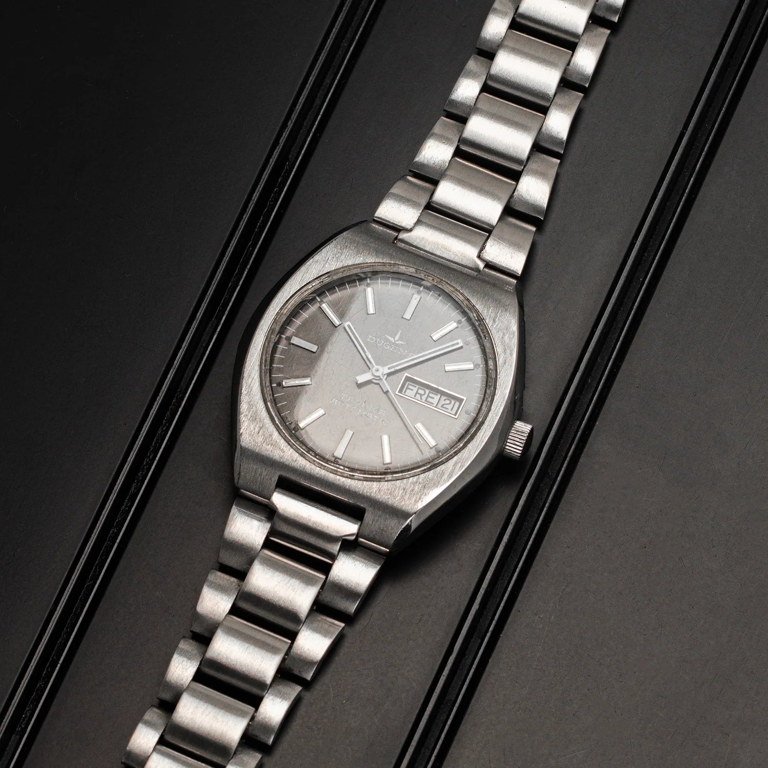 A vintage-style stainless steel wristwatch Dugena Tropica with a textured silver dial and matching metal link bracelet, displayed diagonally on a dark textured background.
