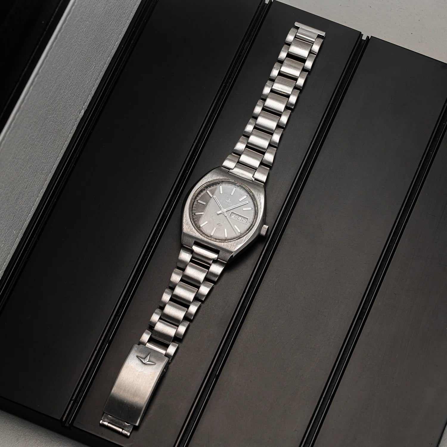 A silver-toned metal wristwatch Dugena Tropica with a matching link bracelet, displayed diagonally on a black presentation box.