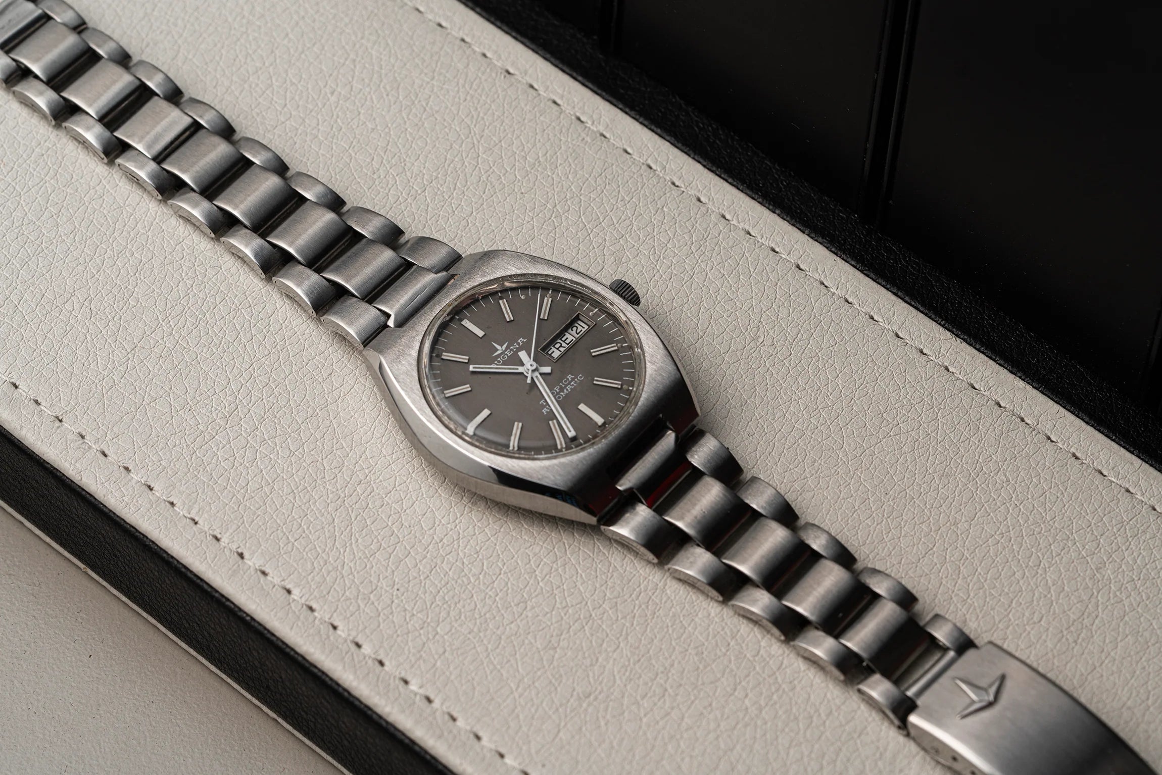 A silver-toned stainless steel wristwatch Dugena Tropica with a dark gray dial and matching metal link bracelet, resting diagonally on a light beige textured leather surface.