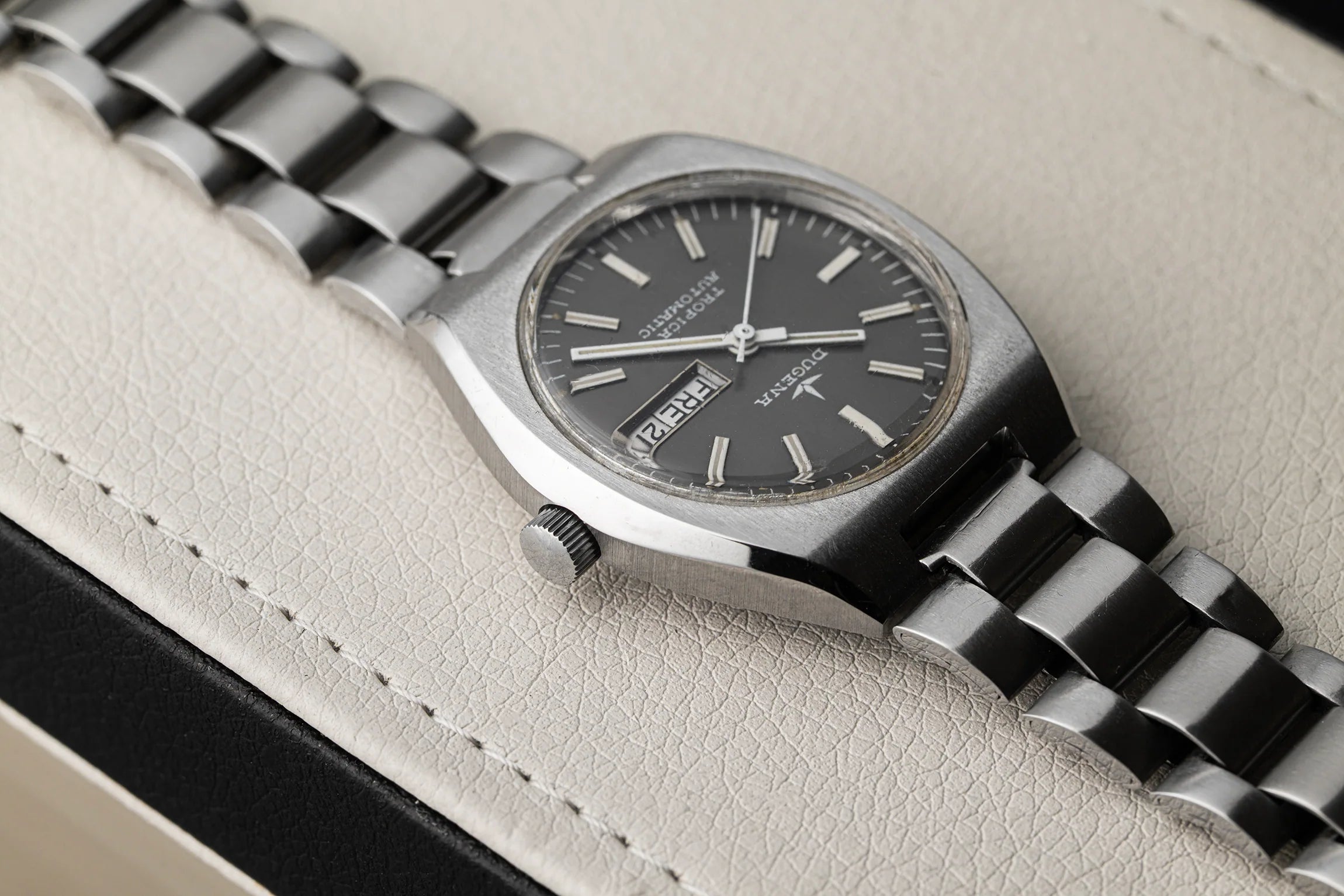 A close-up of a vintage Dugena Tropica wristwatch with a silver-tone metal bracelet and a dark gray dial, resting on a textured light beige leather surface.