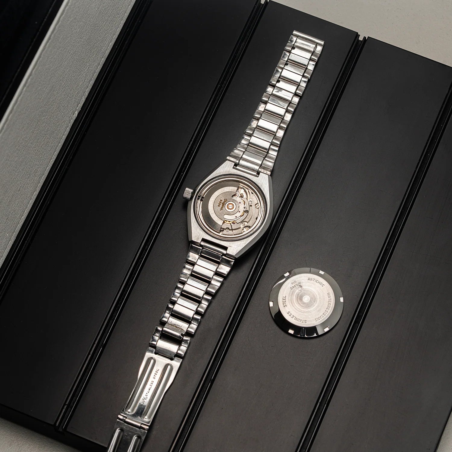 A silver stainless steel automatic watch Dugena Tropica with a visible mechanical movement through the transparent back case, displayed on a black presentation box next to its circular backplate.