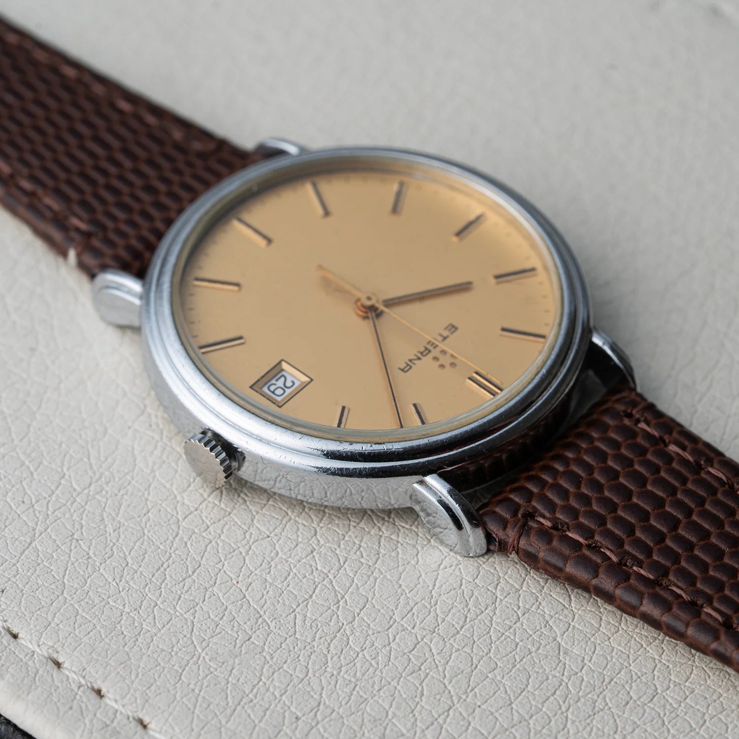 A vintage-style wristwatch Eterna Quartz, Cal. ETA 955.112 with a silver-toned metal case, a warm beige dial, and a textured brown leather strap, resting on a light beige textured surface.