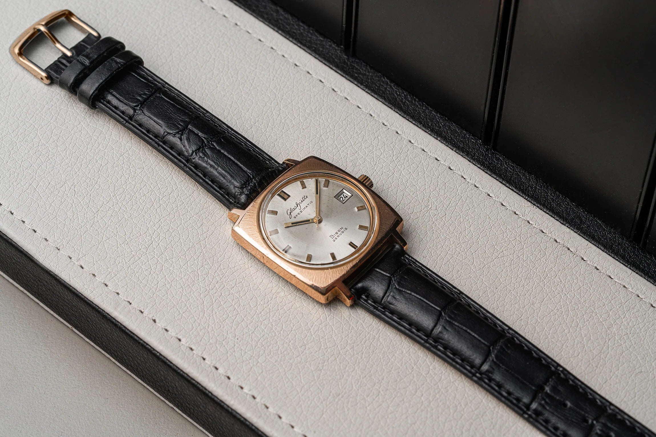 A vintage-style wristwatch Glashutte Bison with a rectangular rose gold case, white dial, and black crocodile-embossed leather strap, resting diagonally on a light gray textured surface.
