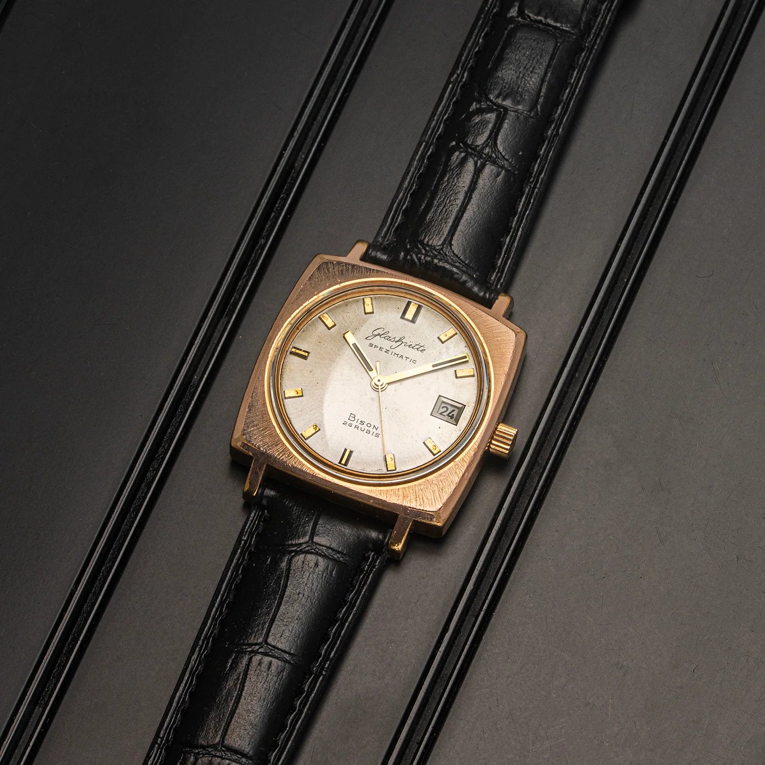 A vintage Glashutte Bison watch with a square rose gold case, a white dial with gold markers, and a black alligator leather strap, displayed diagonally on a dark textured background.