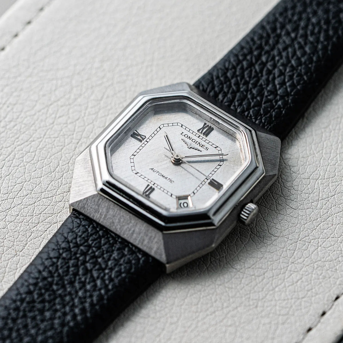 A close-up of a LLongines Octagon, Ref. 4817-4, Cal. L633.1, Men’s Automatic, 1976watch with an octagonal stainless steel case, silver dial, and textured black leather strap, resting on a light gray textured surface.
