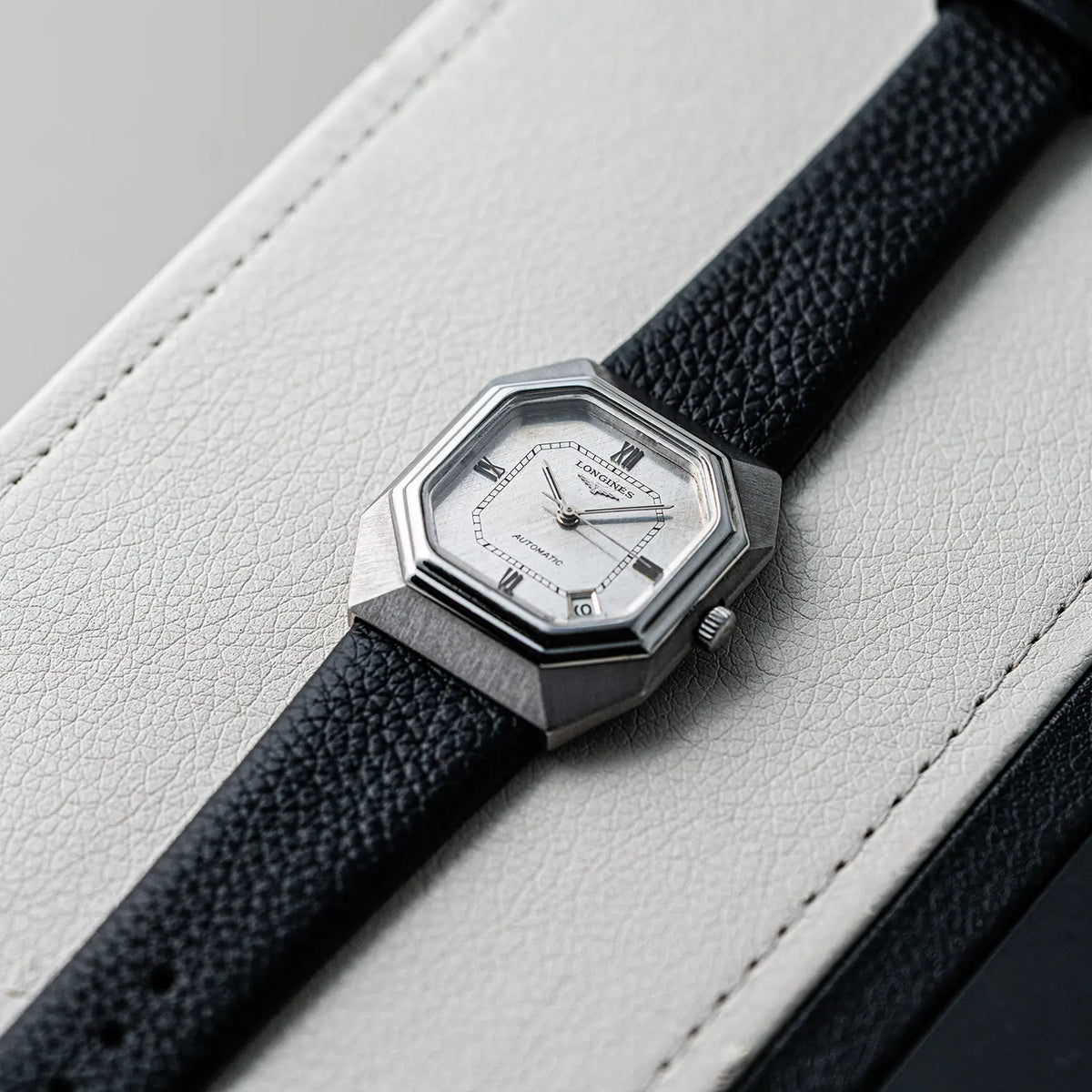 A close-up of a vintage-style wristwatch Longines Octagon, Ref. 4817-4, Cal. L633.1, Men’s Automatic, 1976with an octagonal silver-toned case, white dial, and textured black leather strap, resting diagonally on a white leather surface.
