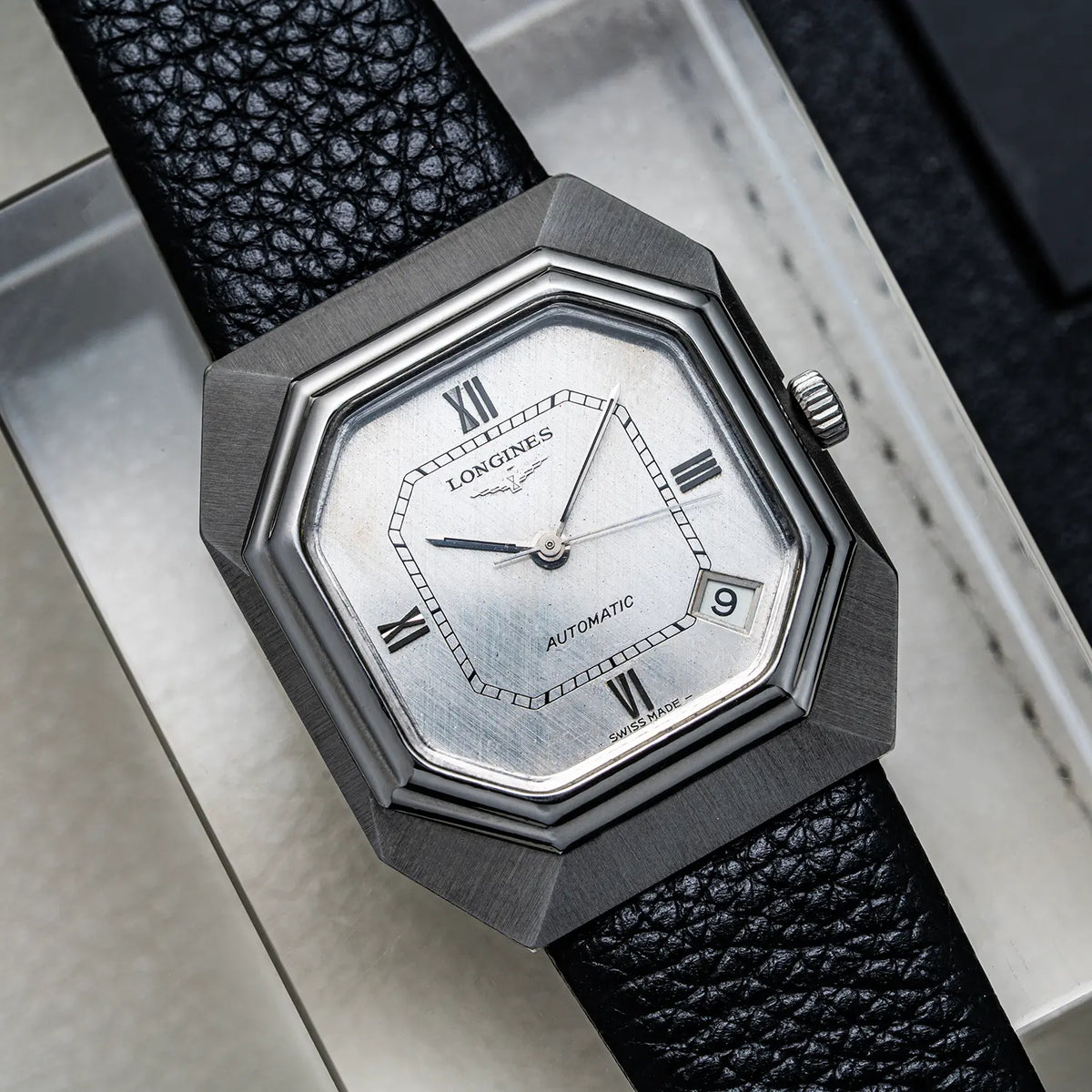 A Longines Automatic watch with an octagonal brushed stainless steel case, a silver dial with Roman numerals, and a textured black leather strap, resting on a metallic surface.
