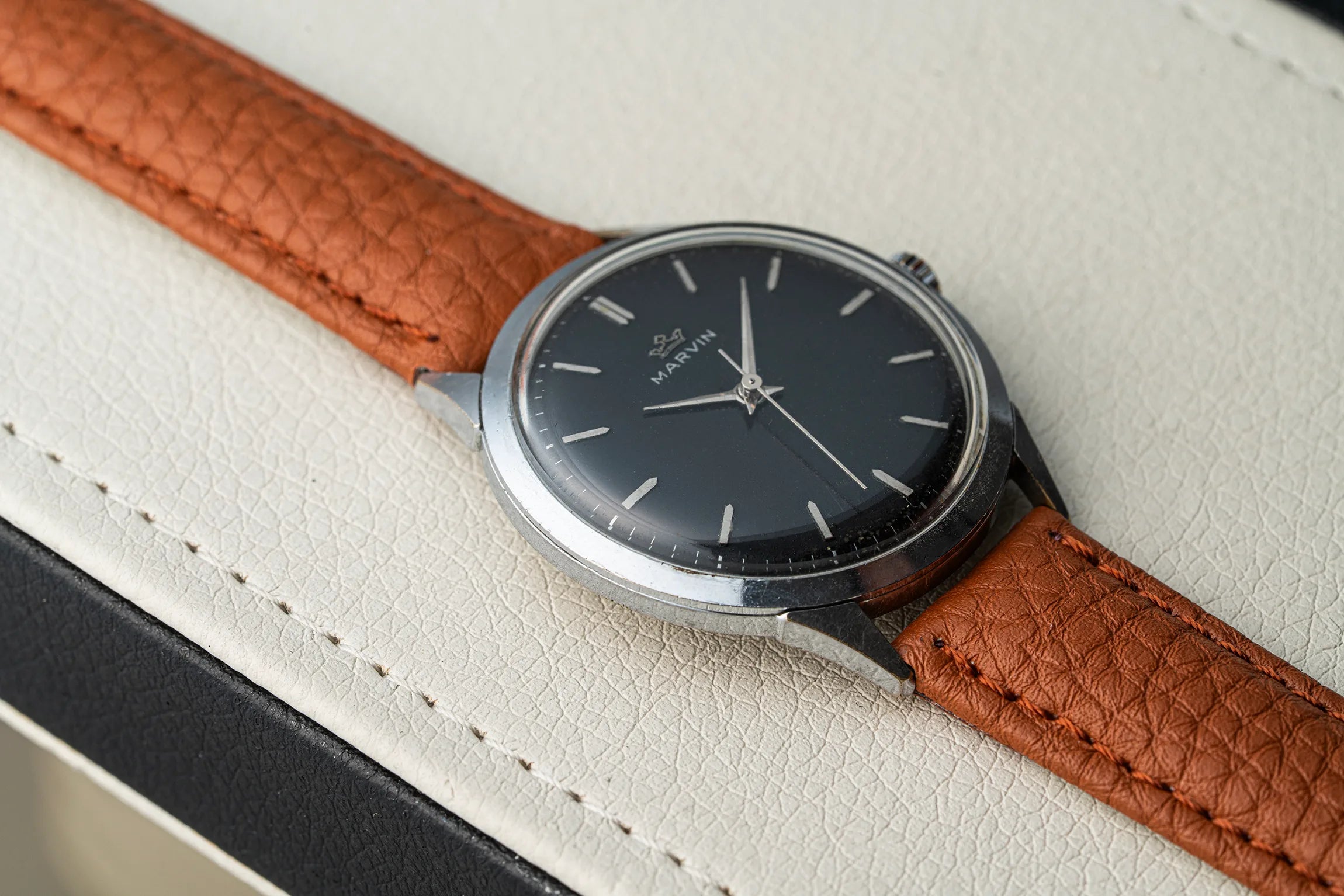 A vintage-style wristwatch Marvin Cal. 560 with a dark charcoal dial, silver-toned case, and a textured brown leather strap, resting on a light cream textured surface.