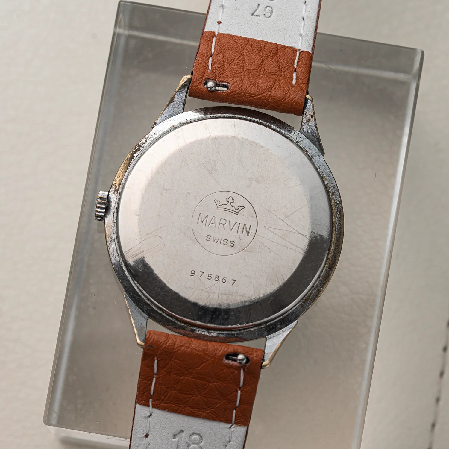 Close-up of the back of a vintage wristwatch Marvin Cal. 560 with a round brushed silver case and a brown leather strap, displayed on a clear acrylic stand. The case is engraved with 'MARVIN SWISS'.