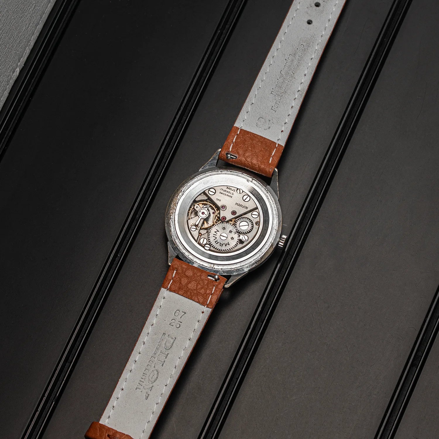 A vintage-style Marvin Cal. 560 watch with a silver-toned case, visible mechanical movement, and a light gray strap with brown leather ends, laid diagonally on a dark textured surface.