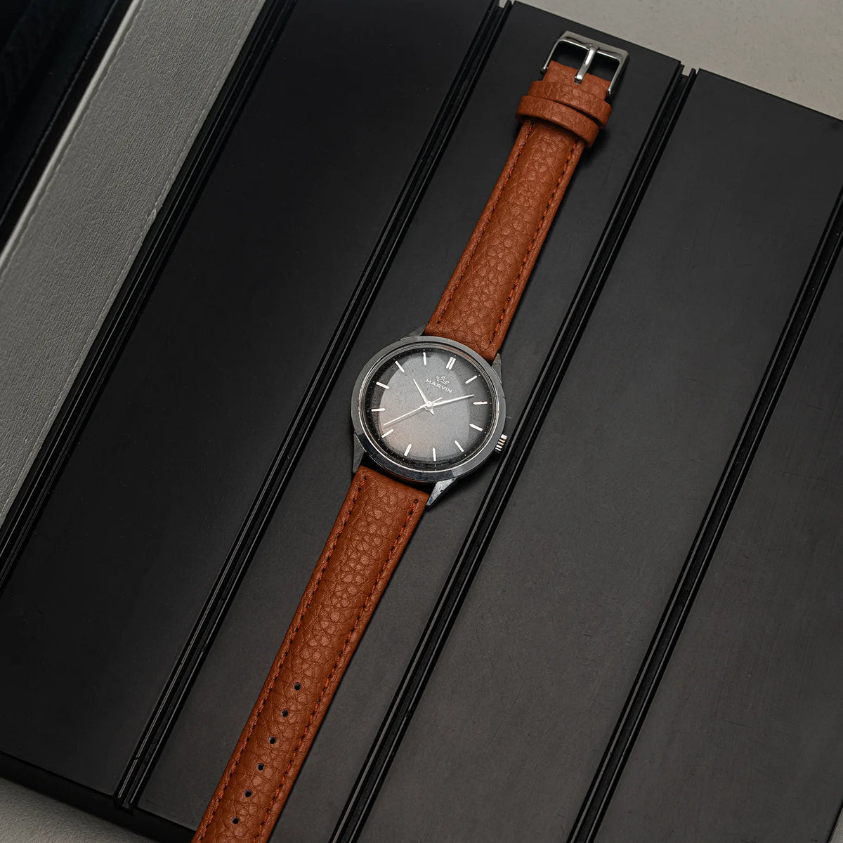 A wristwatch Marvin Cal. 560 with a brown textured leather strap and a dark gray dial, presented diagonally on a black textured background.