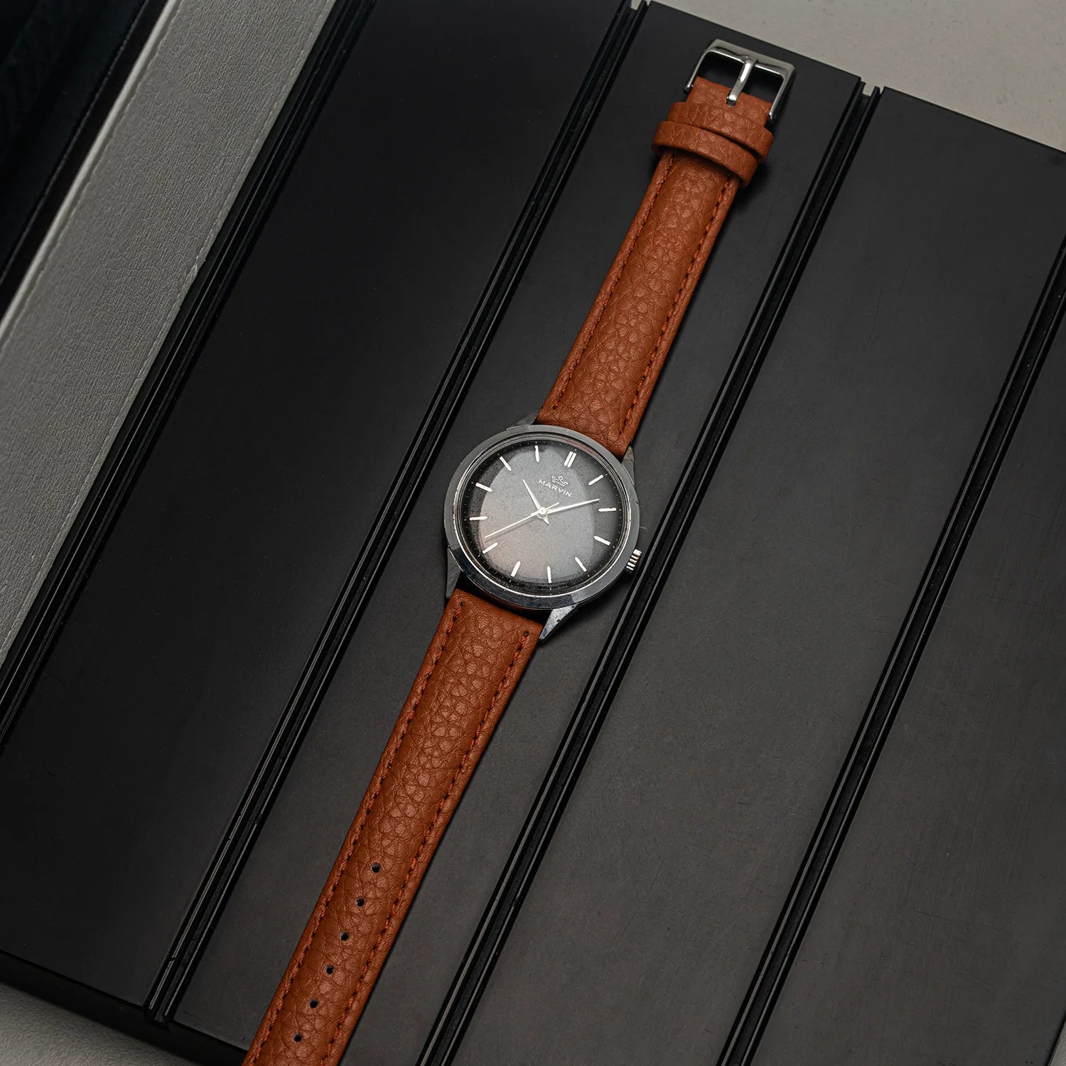 A wristwatch Marvin Cal. 560 with a brown textured leather strap and a dark gray dial, presented diagonally on a black textured background.