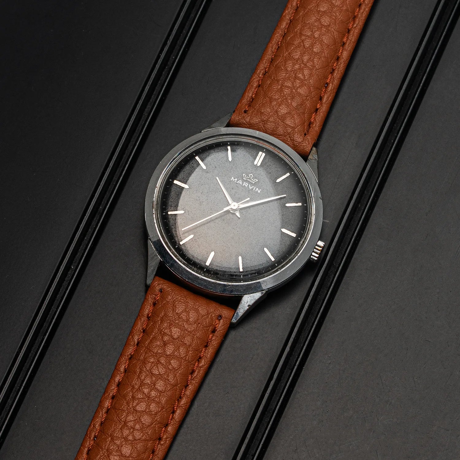 A vintage-style wristwatch Marvin Cal. 560 with a brown textured leather strap and a round silver-tone case, photographed diagonally on a dark gray background with black parallel lines.