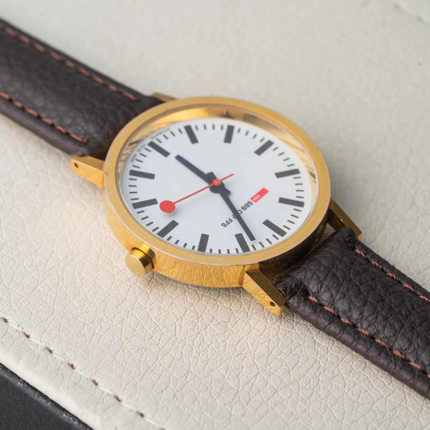 A close-up of a wristwatch Mondaine Official Swiss Railways Watch with a gold-tone case, white dial, black hour markers and hands, a red second hand and red dot detail, and a dark brown leather strap with orange stitching, resting on a textured light beige surface.