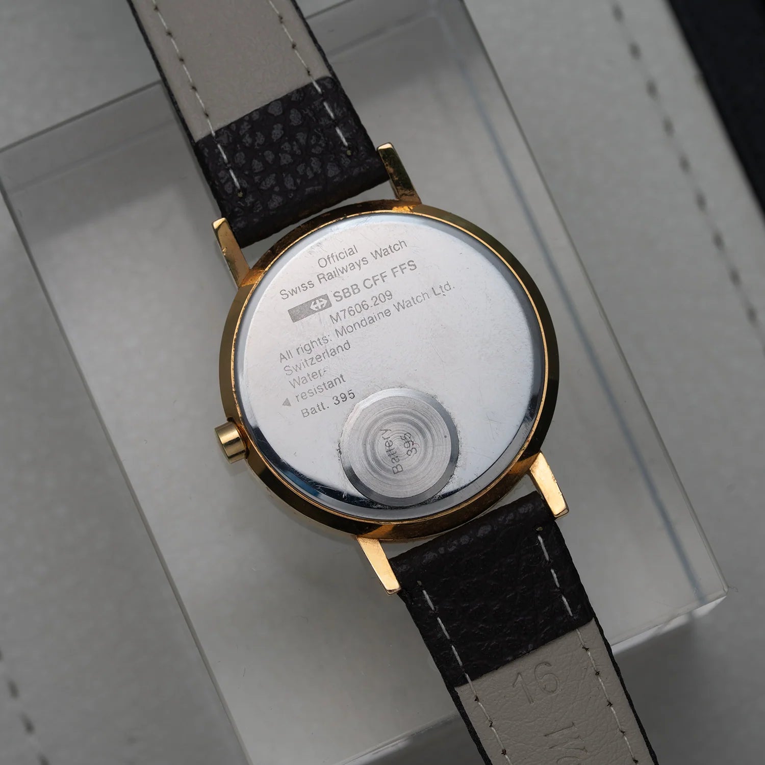 Back view of a wristwatch Mondaine Official Swiss Railways Watch with a gold-toned case and a dark textured strap, resting on a light gray watch box. The metal backplate shows printed specifications.