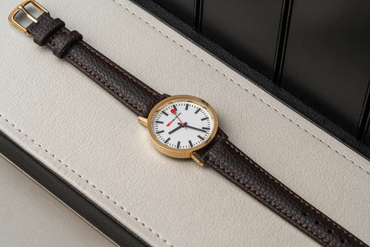 A wristwatch with a gold-tone case, white dial, and dark brown textured leather strap, laid diagonally on a light beige leather surface with a dark background.