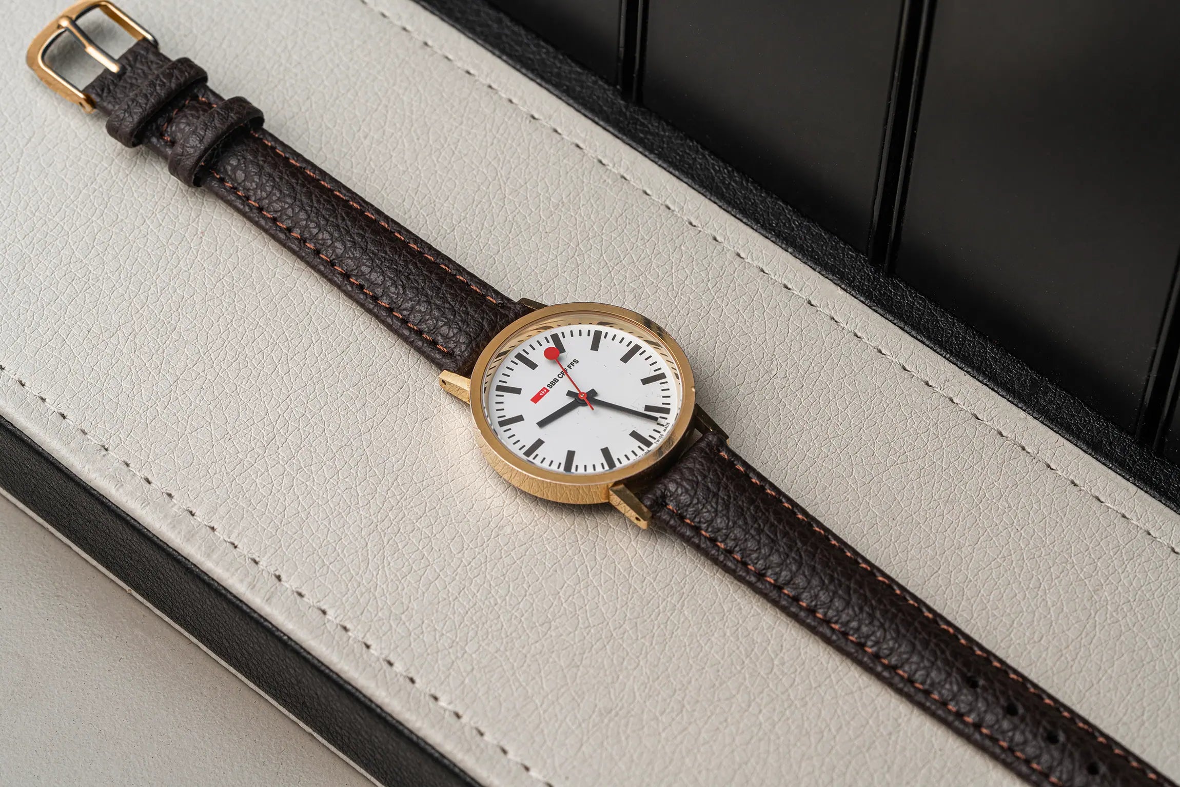 A wristwatch with a gold-tone case, white dial, and dark brown textured leather strap, laid diagonally on a light beige leather surface with a dark background.