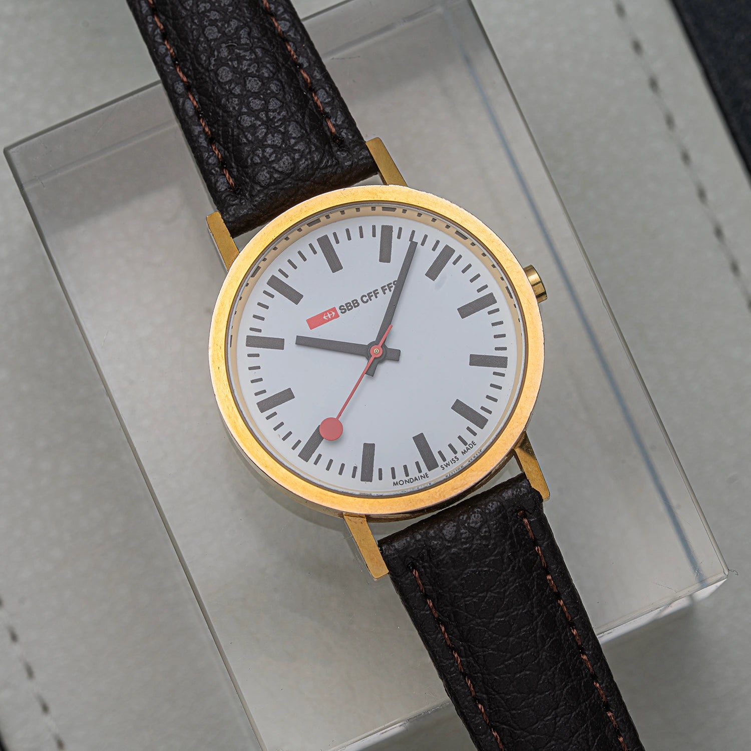 A wristwatch Mondaine Official Swiss Railways Watch with a round gold-tone case, white dial, black hour markers and hands, and a dark brown textured leather strap, displayed on a light gray surface.