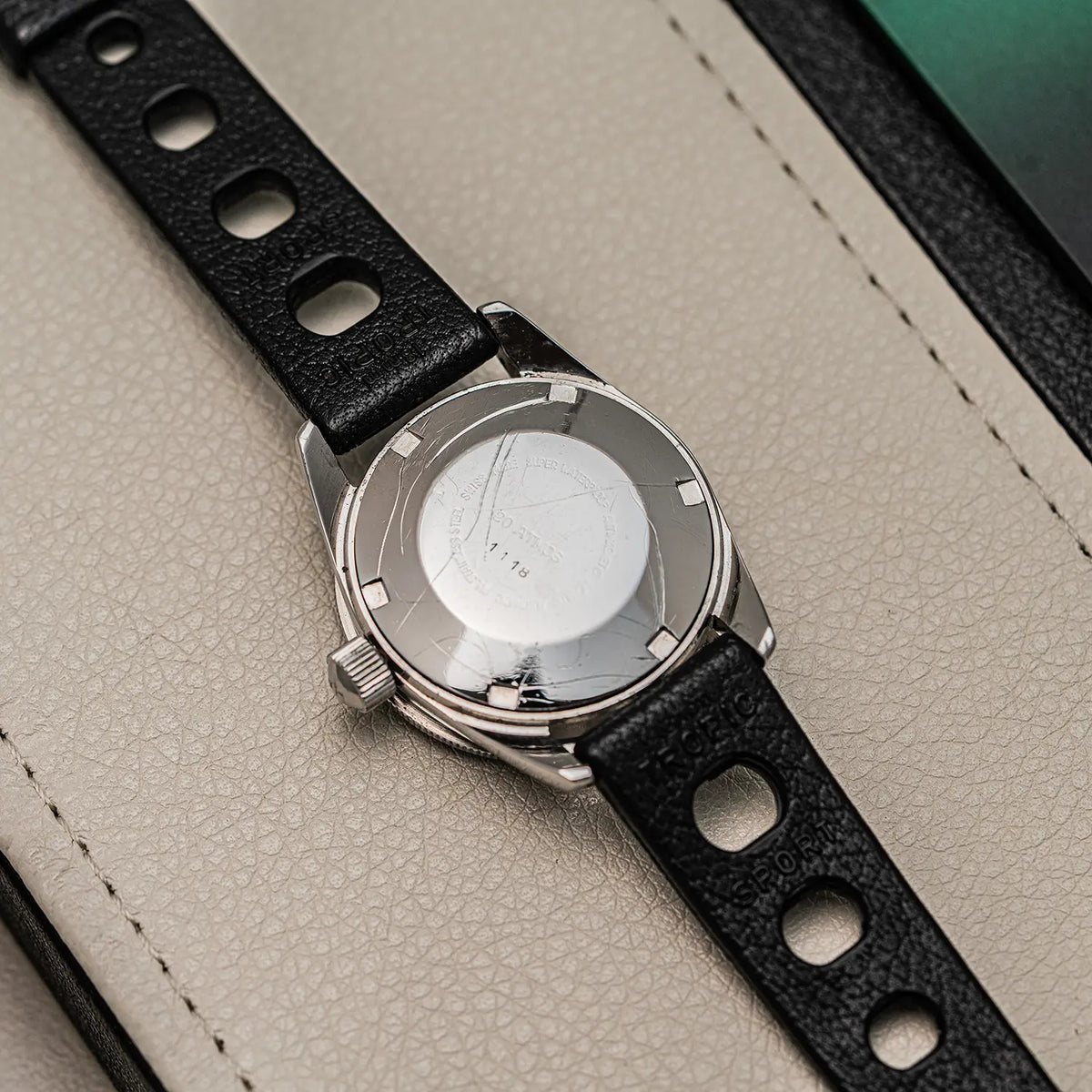 Close-up of a wristwatch with a black perforated leather strap and a silver-toned metal case back, resting on a light beige textured surface.