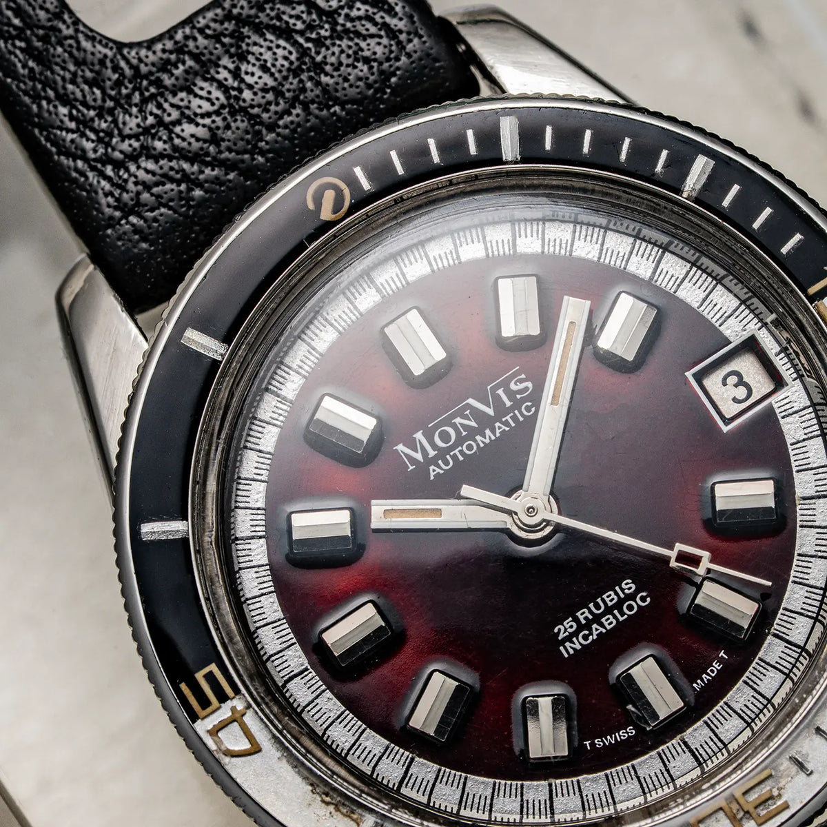 Close-up of a vintage Monvis Bakelite Diver, 20 ATM, Ref. 1.18, Cal. ETA 2783, Men’s Divingwatch with a black leather strap, black bezel, and a dark red gradient dial with silver-tone hour markers.