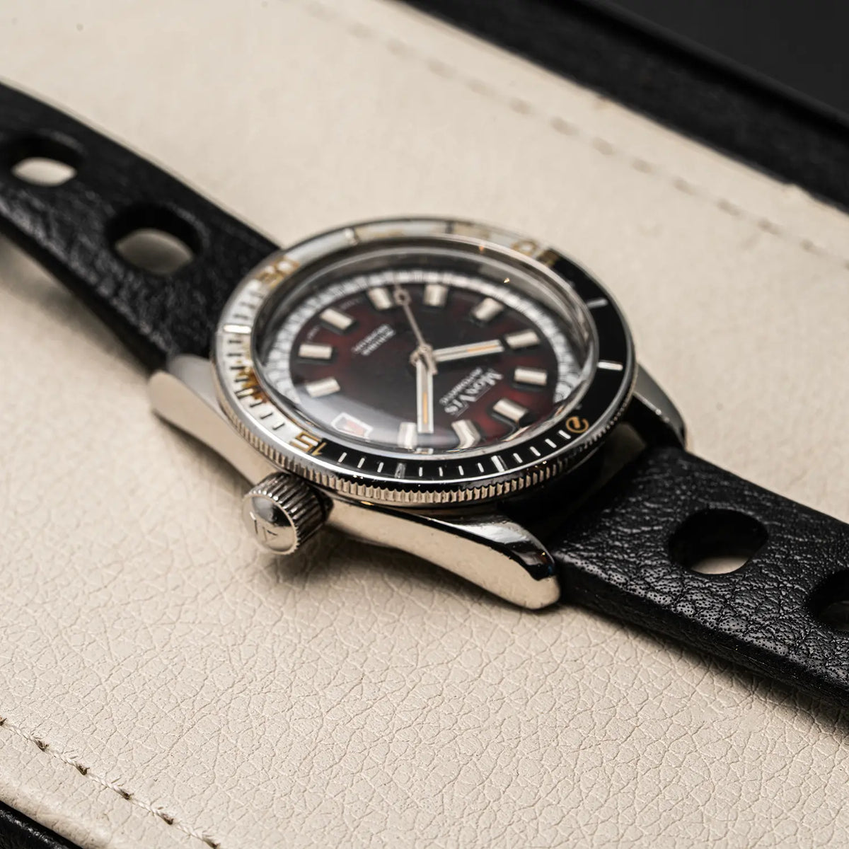 A close-up of a vintage-style wristwatch Monvis Bakelite Diver, 20 ATM, Ref. 1.18, Cal. ETA 2783, Men’s Divingwith a black dial, black bezel, silver-toned case, and black perforated leather strap, resting on a light beige textured surface.