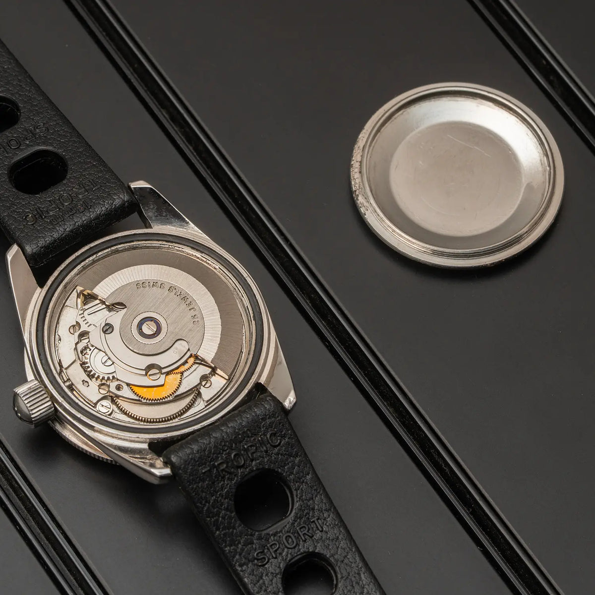 A close-up of a wristwatch with its back cover removed, showing the exposed mechanical movement and black textured strap, placed on a dark surface next to a round silver metal piece.