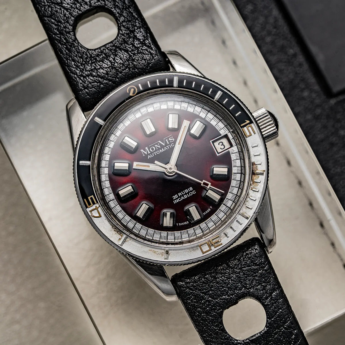A Monvis Bakelite Diver, 20 ATM, Ref. 1.18, Cal. ETA 2783, Men’s Diving wristwatch with a dark red dial, silver-tone stainless steel case, and a textured black leather strap, shown resting on a light-colored surface.
