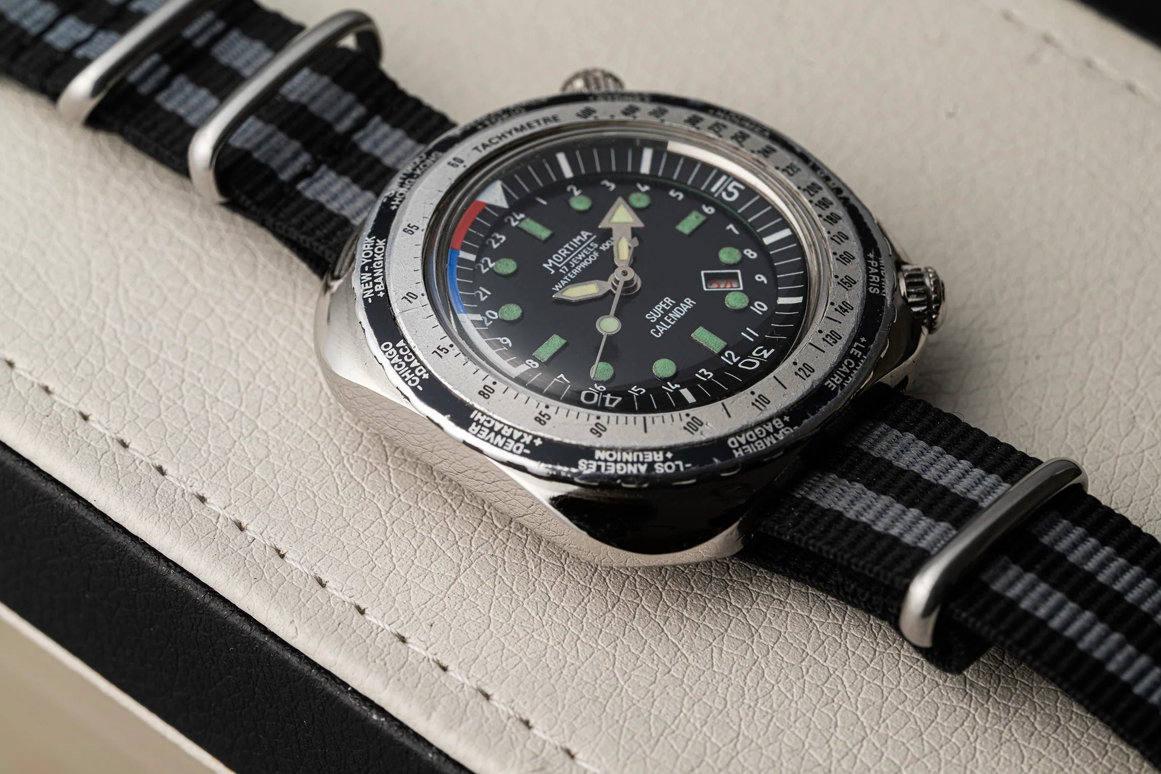 A close-up of a wristwatch Mortima Super Calendar with a black dial, silver-toned case, and black-and-gray striped fabric strap, resting on a light textured surface.