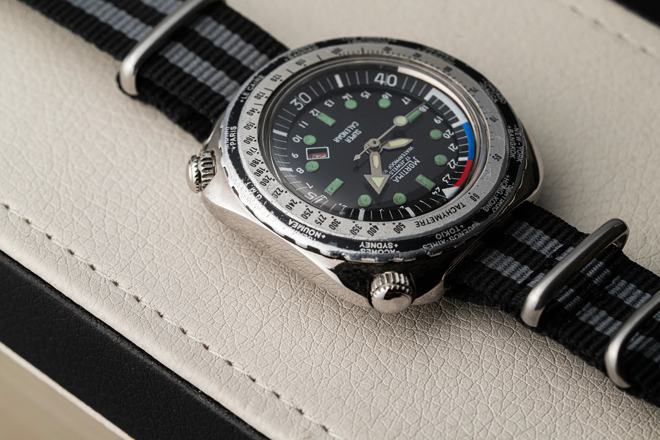 A close-up of a wristwatch Mortima Super Calendar with a black dial, silver-tone case, and black-and-gray striped nylon strap, resting on a light beige textured surface.