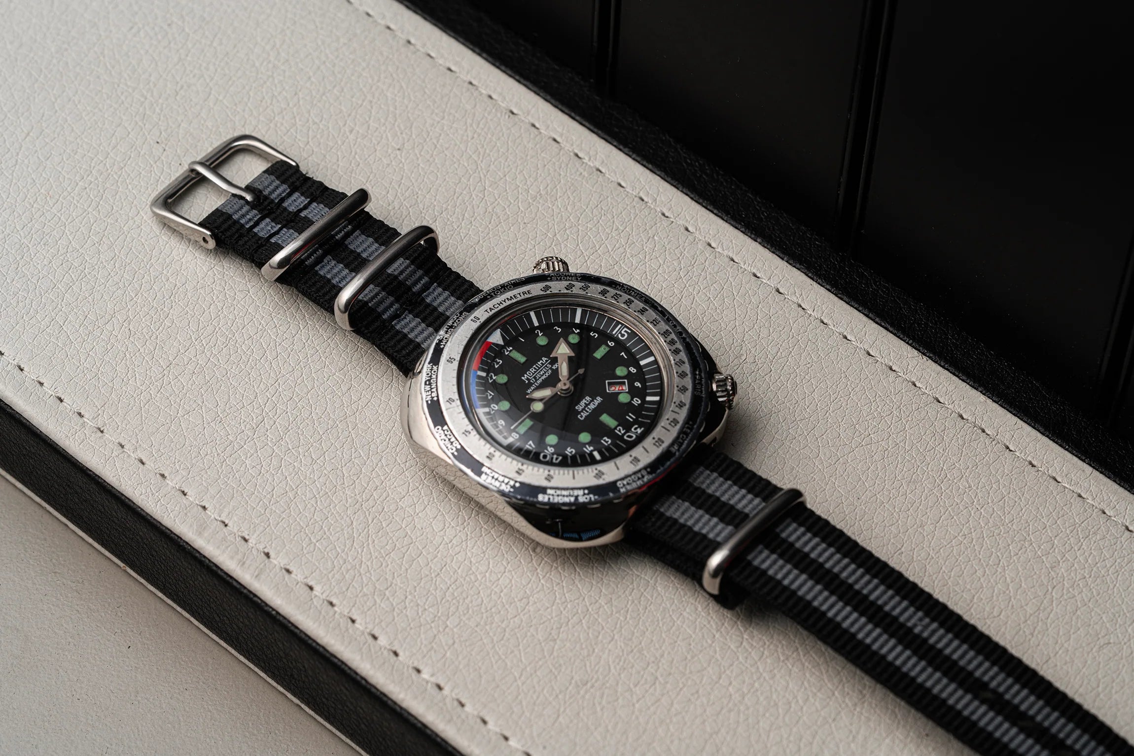 A wristwatch Mortima Super Calendar with a silver-toned case, black dial, and black-and-gray striped nylon strap, resting diagonally on a light beige textured leather surface.
