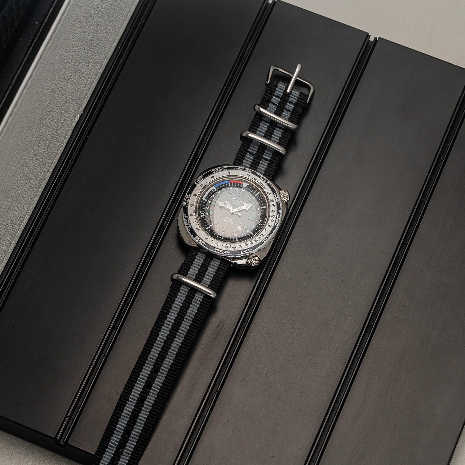 A wristwatch Mortima Super Calendar with a silver-toned case, white dial, and black-and-gray striped nylon strap, displayed diagonally on a dark textured surface.