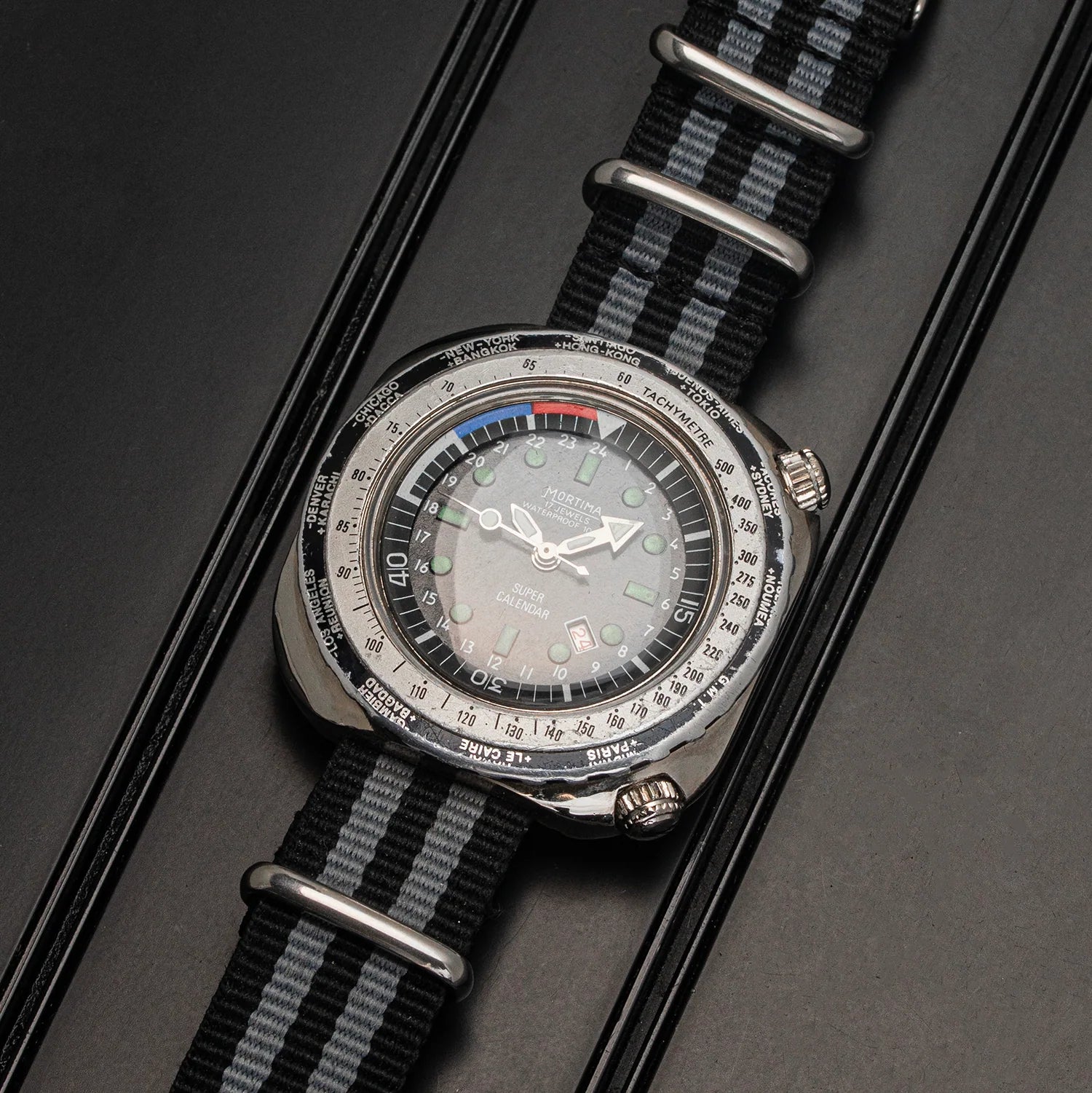 A wristwatch Mortima Super Calendar with a silver-toned case, a gray dial, and a black-and-gray striped nylon strap, photographed at an angle on a dark textured surface.