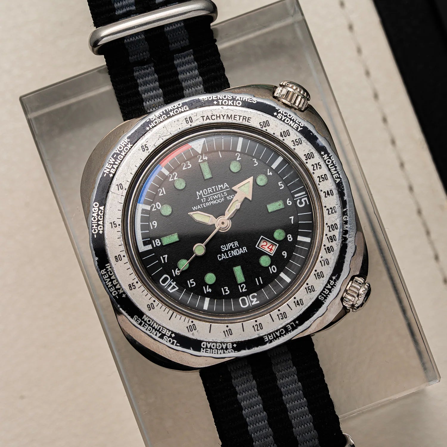 Mortima Super Calendar wristwatch with a black dial, silver-toned case, and black fabric strap, displayed on a clear acrylic stand.