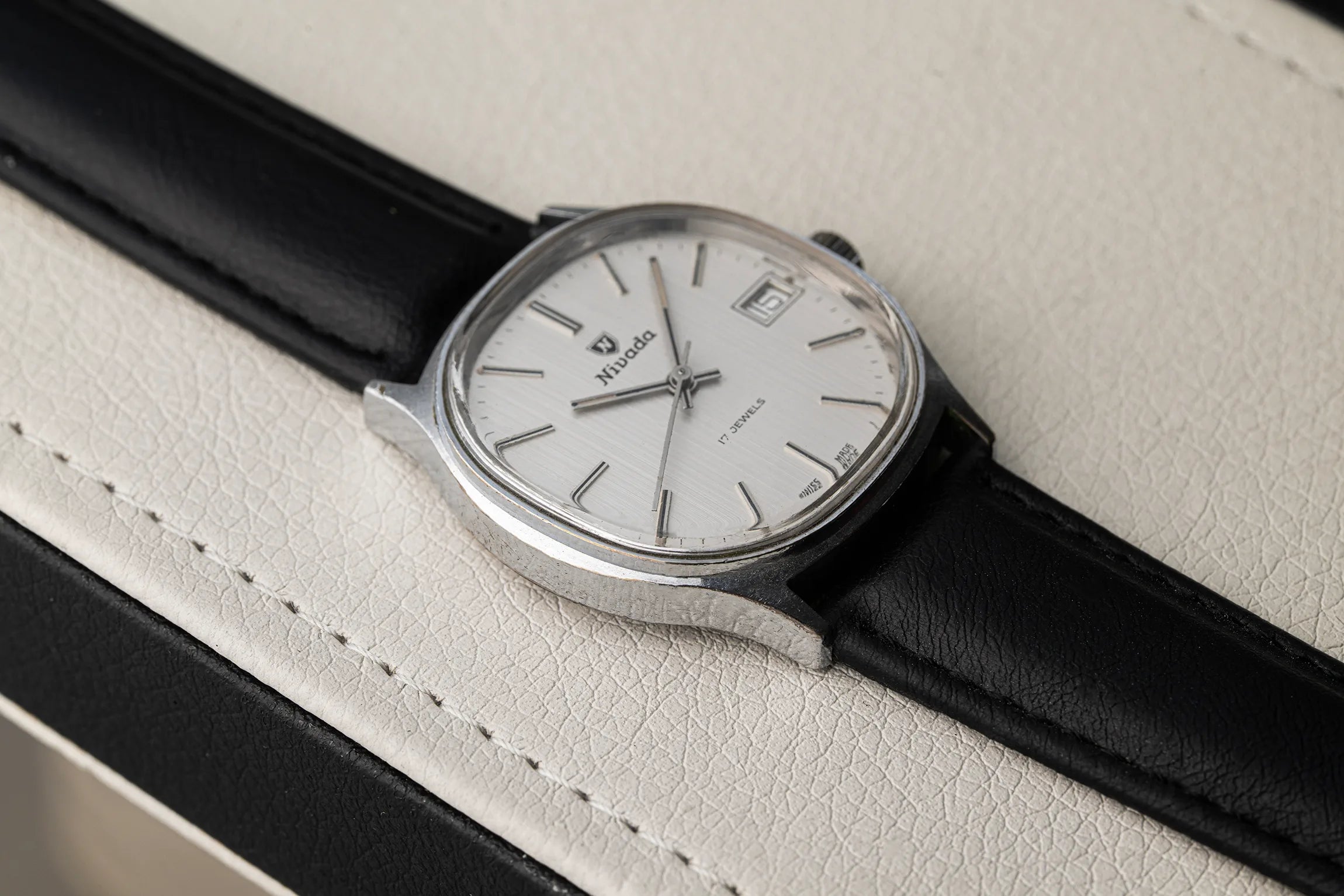 A close-up of a vintage-style wristwatch Nivada Square with a silver-toned case, white dial, and black leather strap, resting on a textured off-white surface.