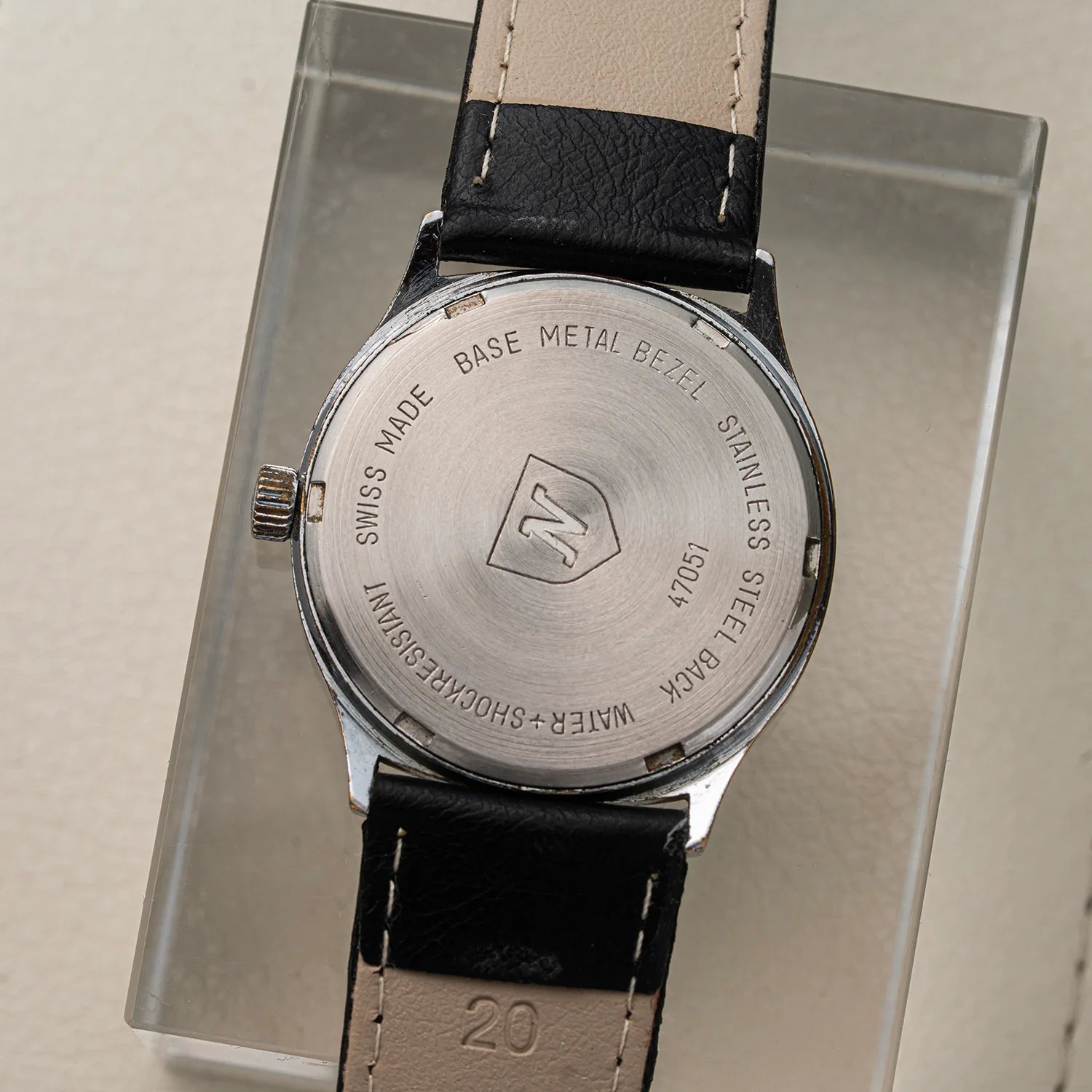 Close-up of the back of a wristwatch Nivada Square with a stainless steel case and black leather strap, resting on a clear display block. The case is engraved with details including 'SWISS MADE' and a central shield logo.