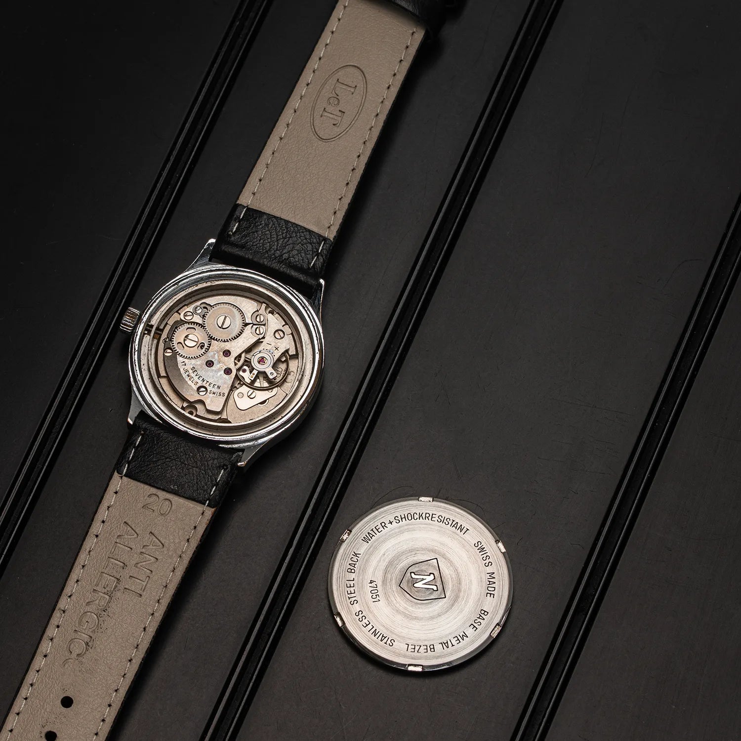 A vintage-style Nivada Square watch with a beige leather strap and silver-toned case, shown open to reveal the mechanical movement, alongside the separate silver case back on a dark textured background.