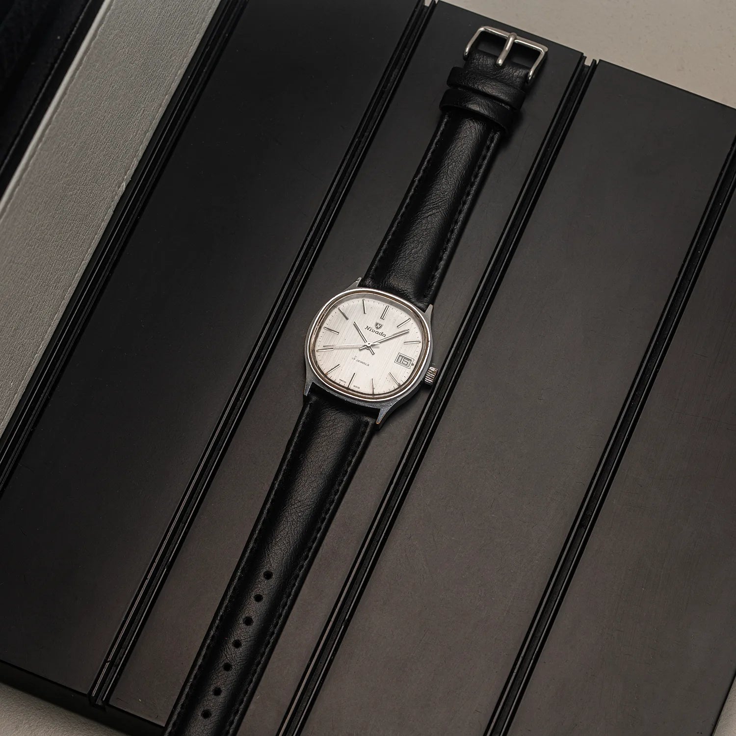 A vintage-style wristwatch Nivada Square with a silver-toned case, white dial, and black leather strap, displayed diagonally on a black textured surface.