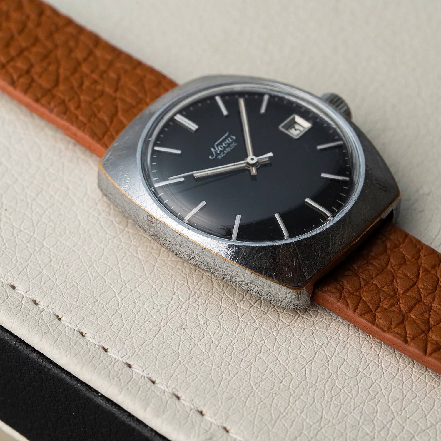 A close-up of a vintage-style wristwatch  Novus, Cal. FE 3612 with a square silver-toned case, black dial, and brown textured leather strap, resting on a light cream textured surface.