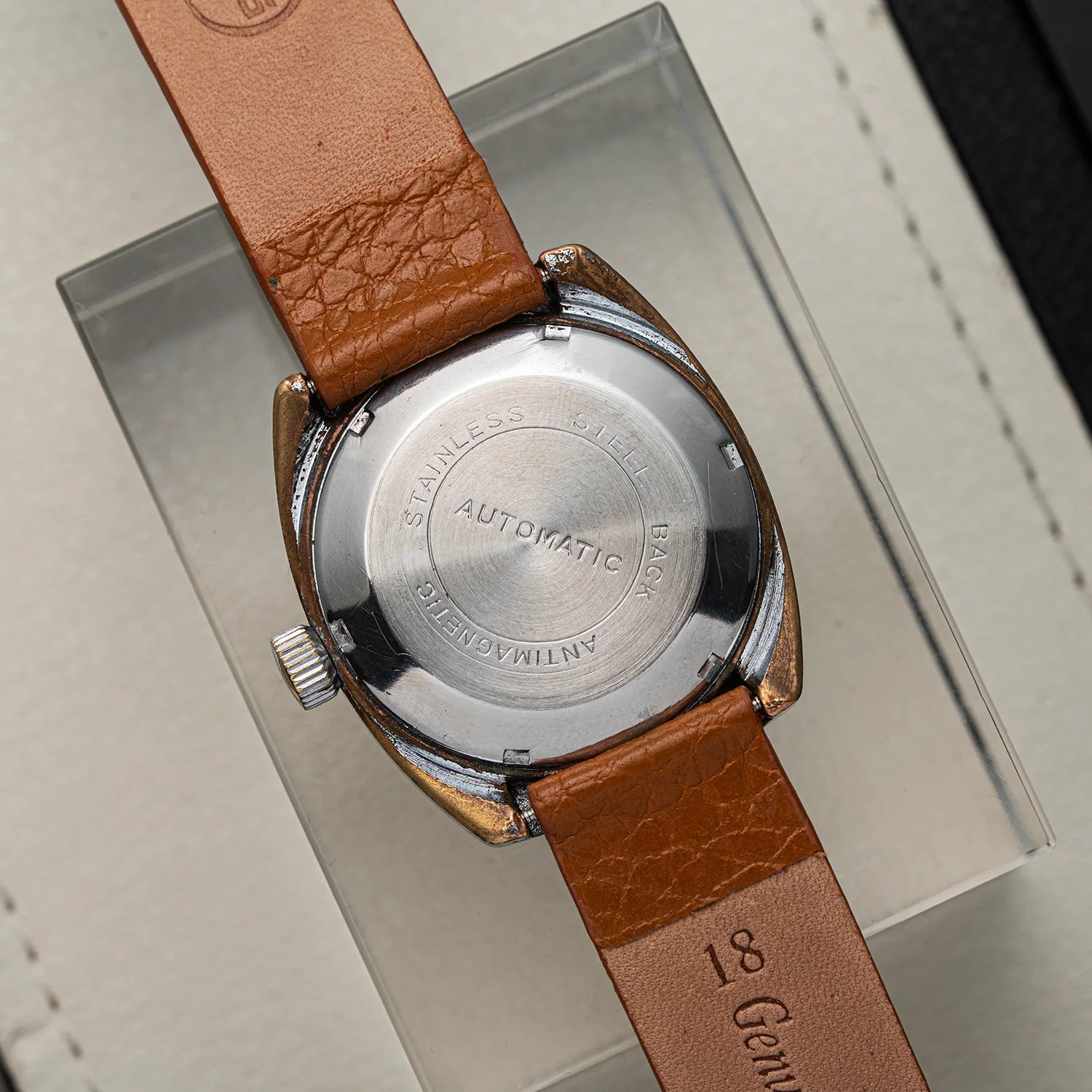 Close-up of the back of a vintage wristwatch with a brown leather strap and a polished stainless steel case, resting on a clear display box.