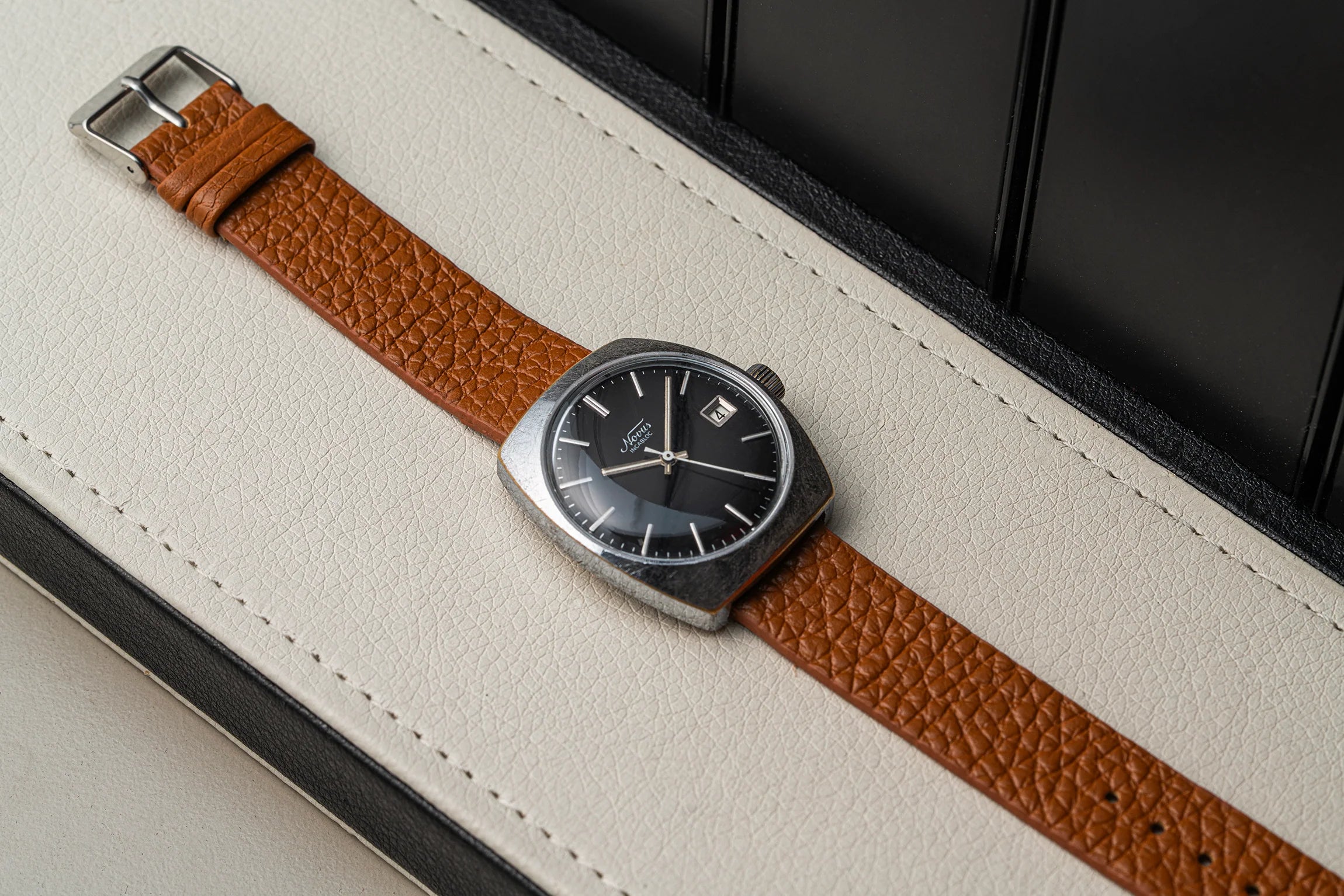 A vintage-style wristwatch Novus, Cal. FE 3612 with a brown textured leather strap and a silver-toned rectangular case, resting diagonally on a cream-colored leather surface.
