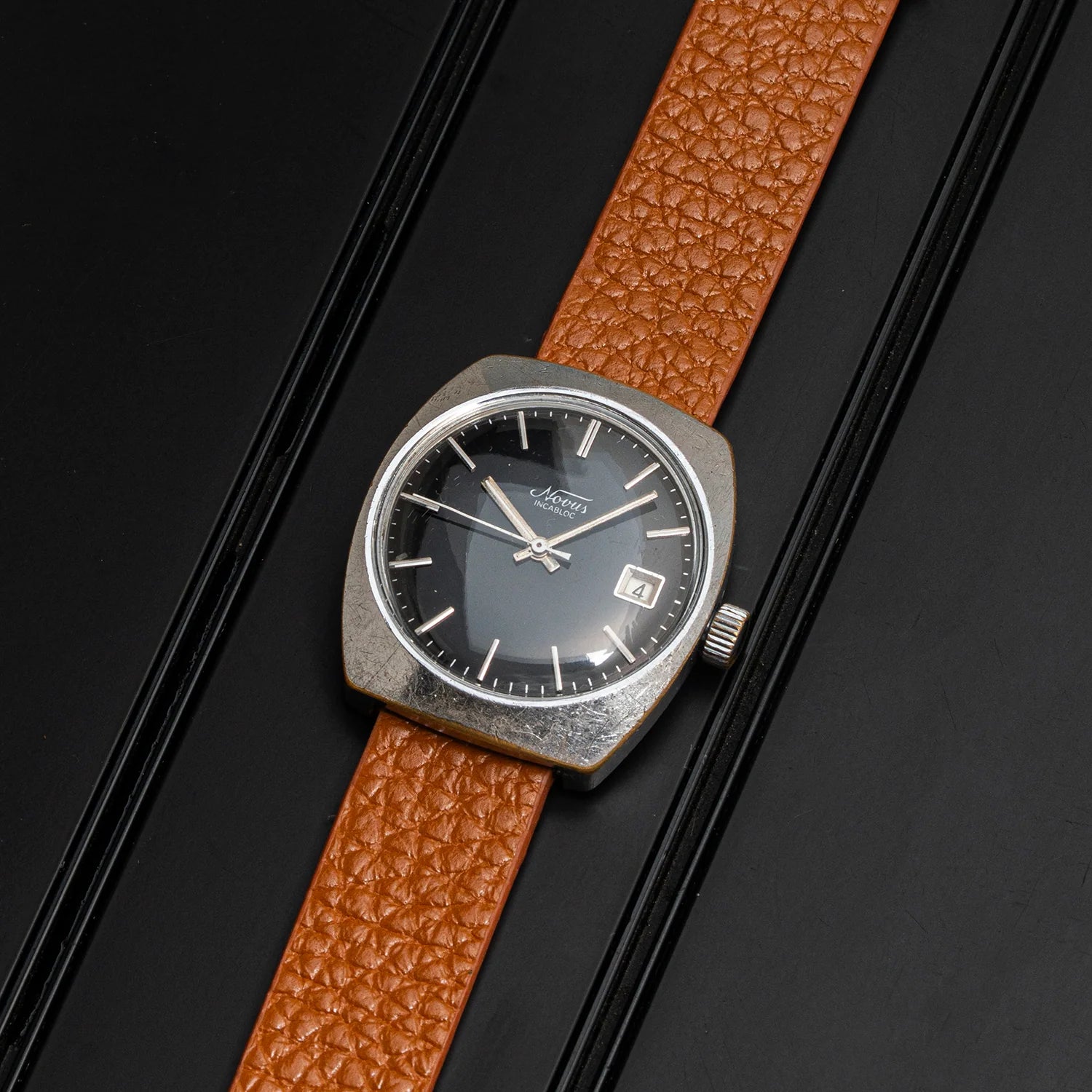 A vintageNovus, Cal. FE 3612 watch with a rectangular silver-toned case, black dial, and textured brown leather strap, displayed diagonally on a black background.