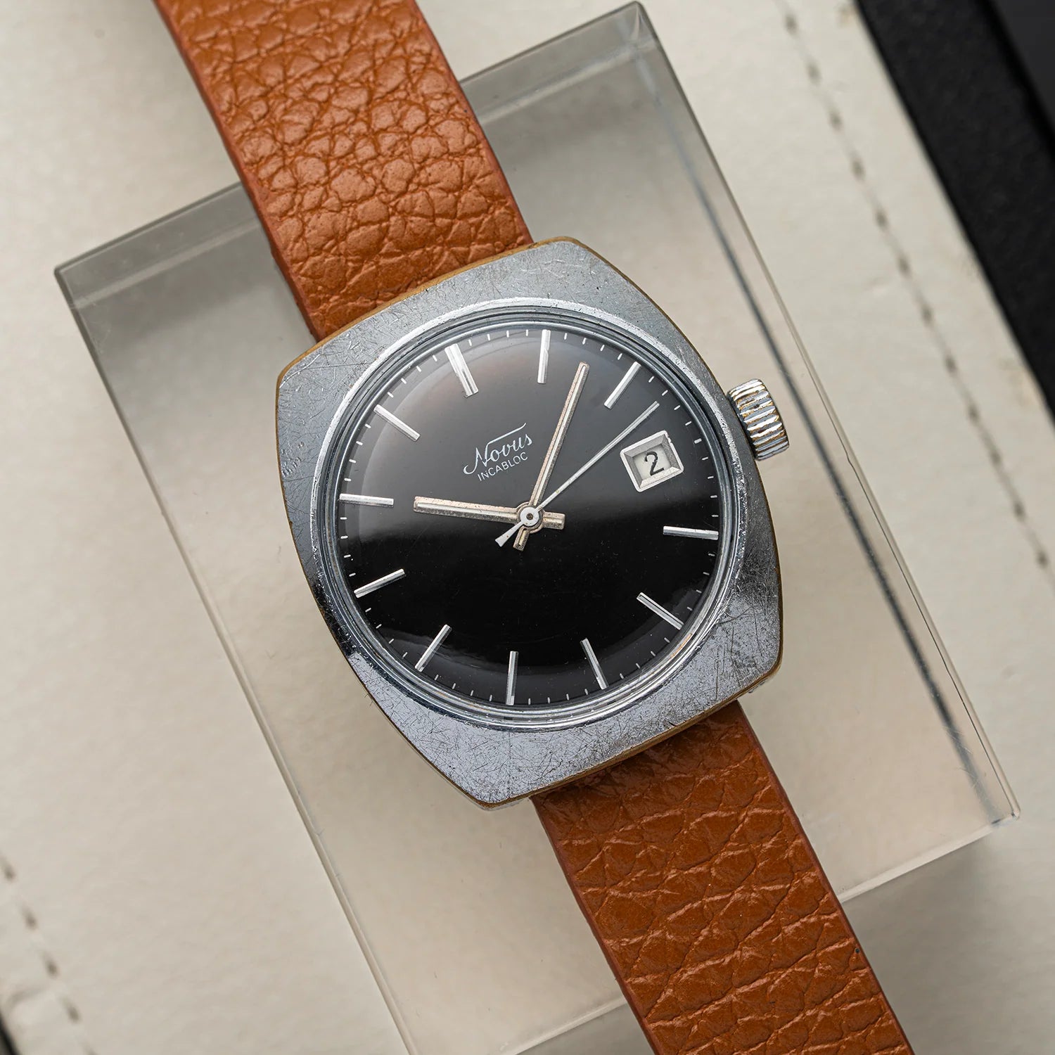 A vintage-style wristwatch Novus, Cal. FE 3612 with a round silver-tone case, black dial, and brown textured leather strap, displayed diagonally on a clear acrylic stand over a light beige surface.